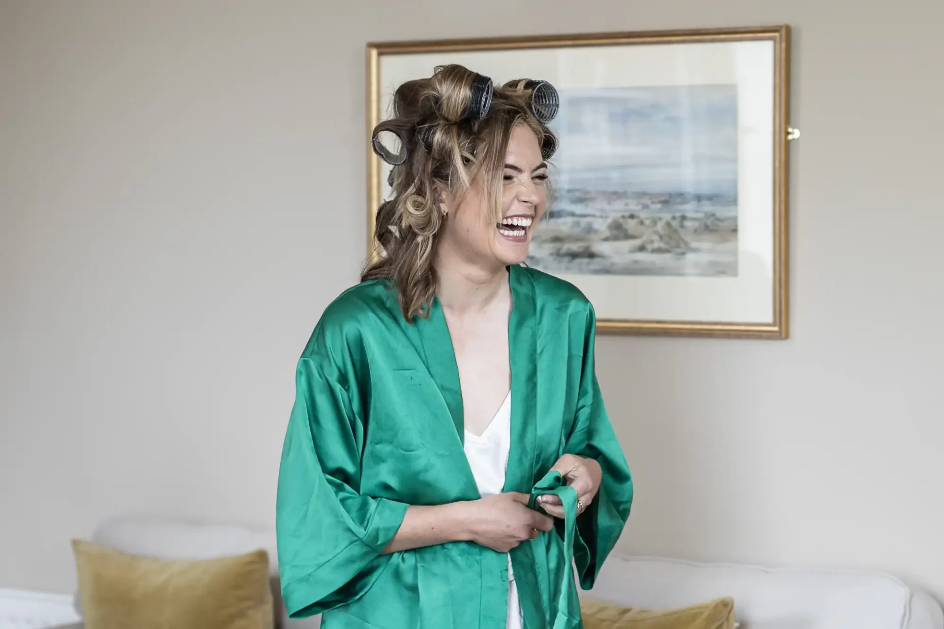 A woman with curlers in her hair, wearing a green robe, smiles broadly in a room with a framed seascape painting on the wall behind her.