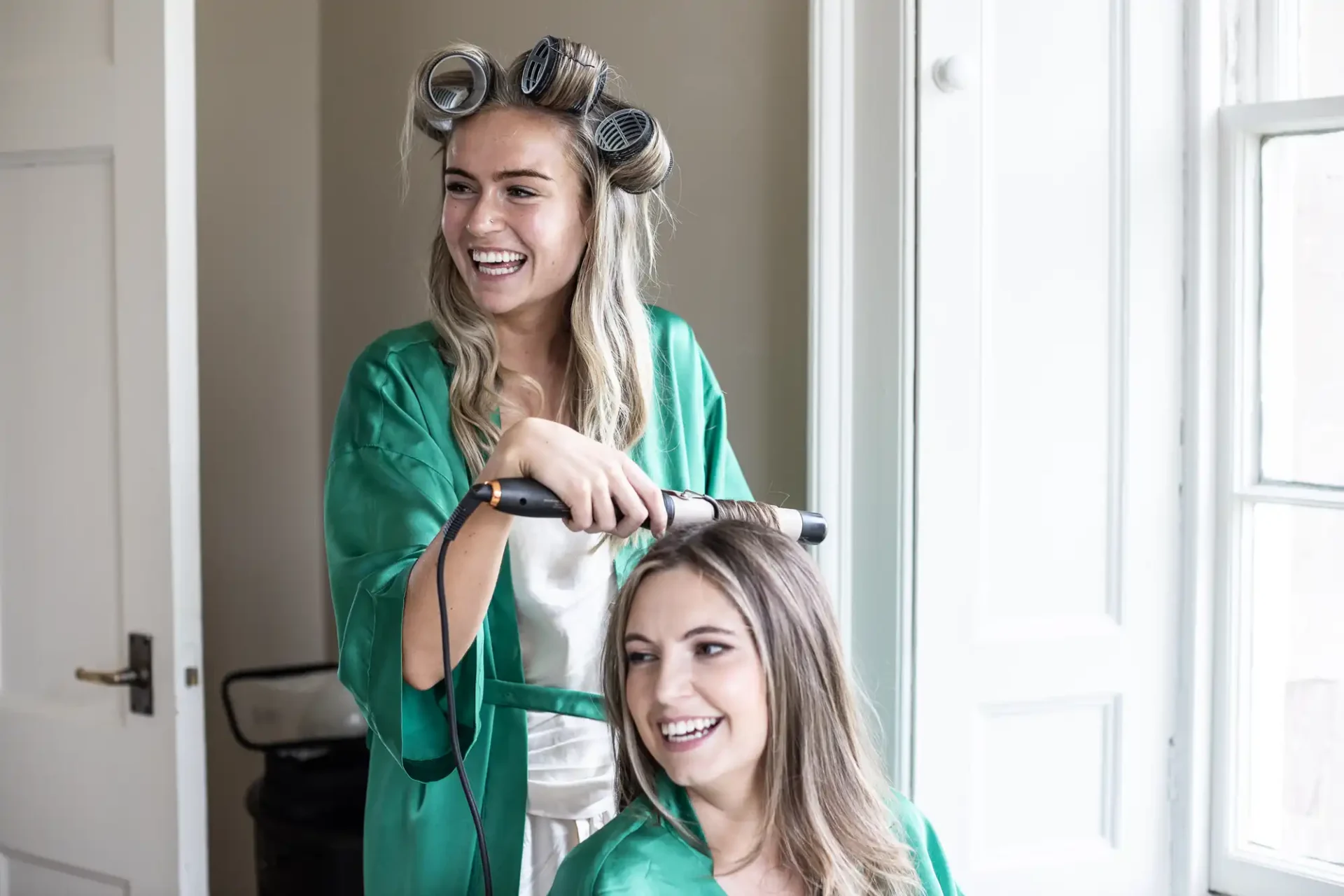 Two women smiling, one standing with hair curlers and holding a curling iron, and the other sitting with straight hair, both wearing green robes.