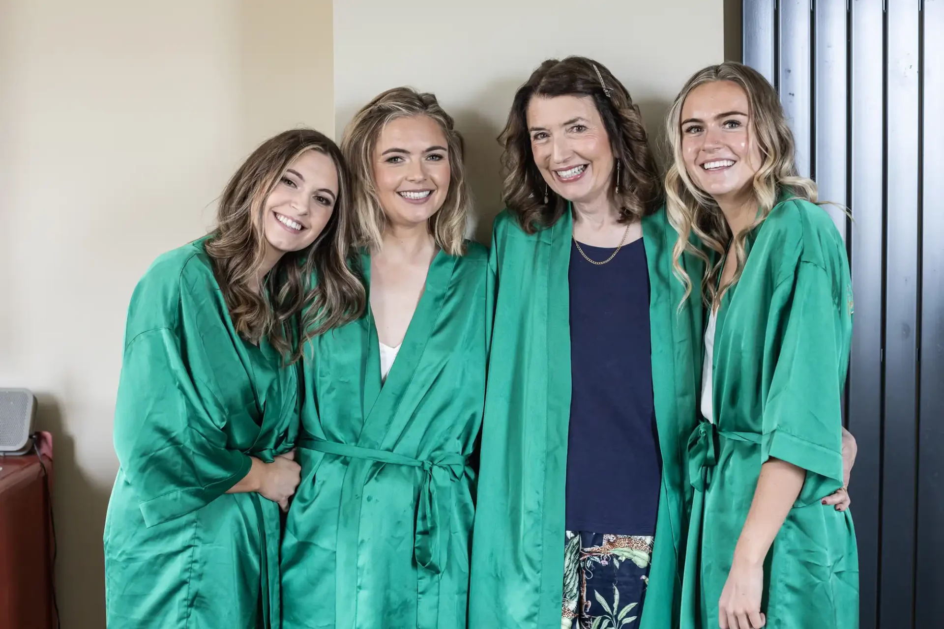 Four women wearing green robes stand close together, smiling, in an indoor setting with light-colored walls and a black vertical panel.