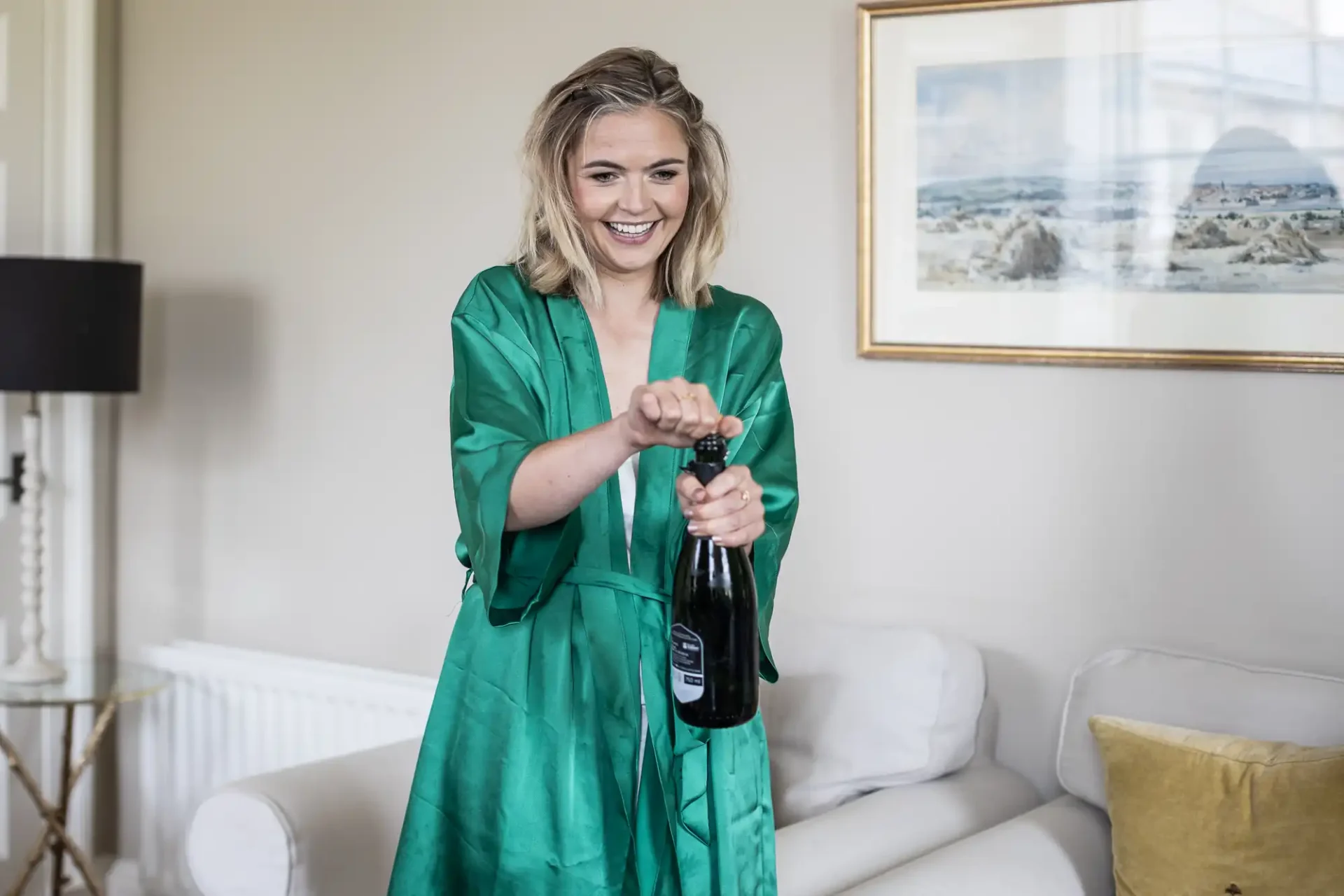 A woman in a green robe is smiling as she opens a bottle of champagne in a living room. A couch and a framed picture are in the background.