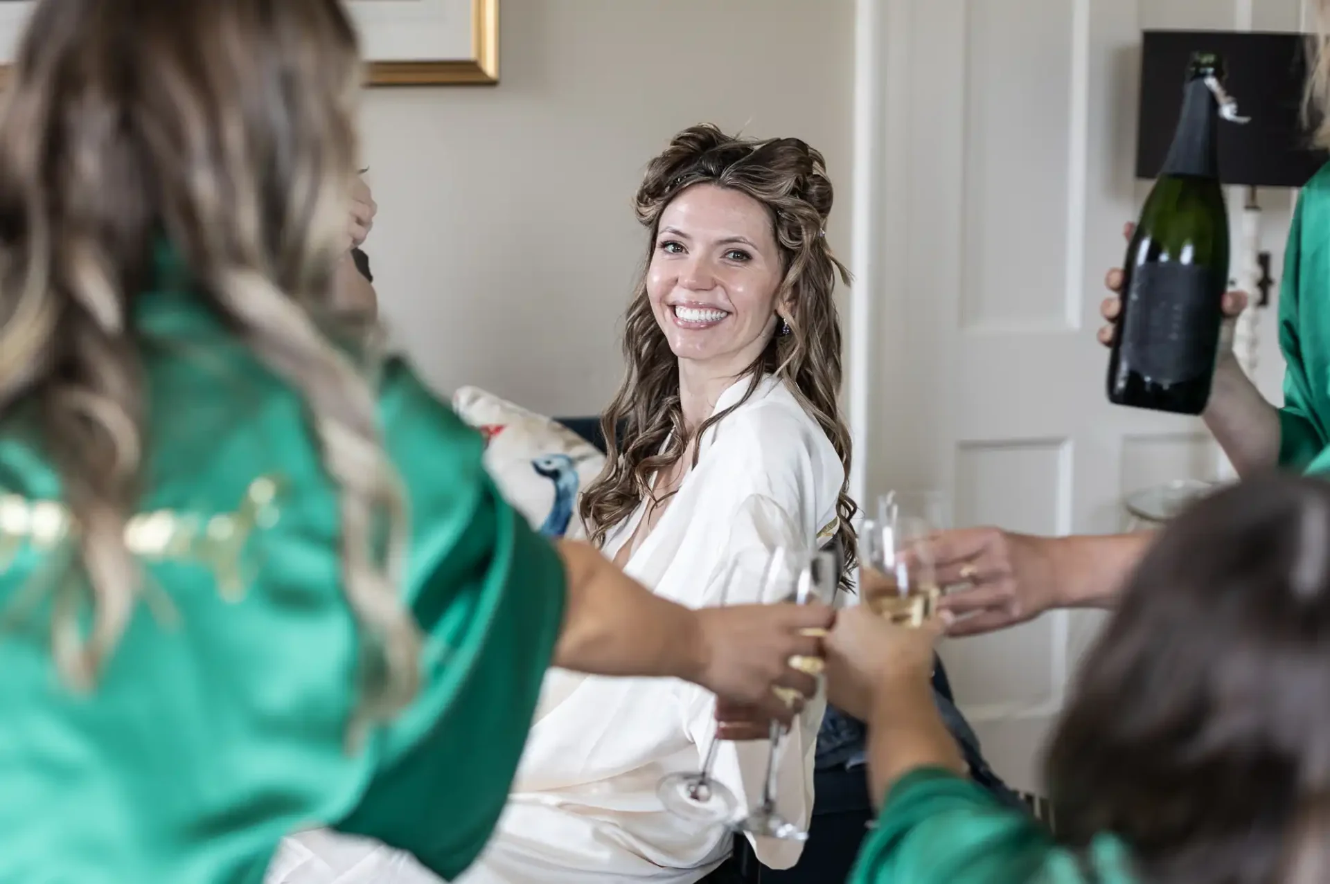 A woman in a white robe smiles as she sits, surrounded by people in green robes holding champagne glasses, about to make a toast.
