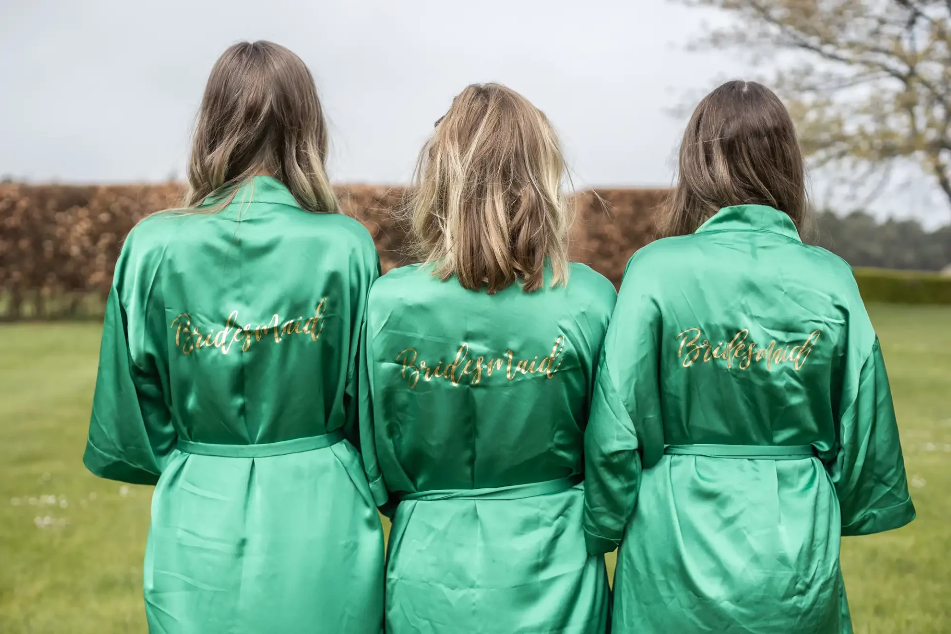 Three women are standing outdoors with their backs to the camera, wearing green robes that have the word "Bridesmaid" embroidered in gold on the back.