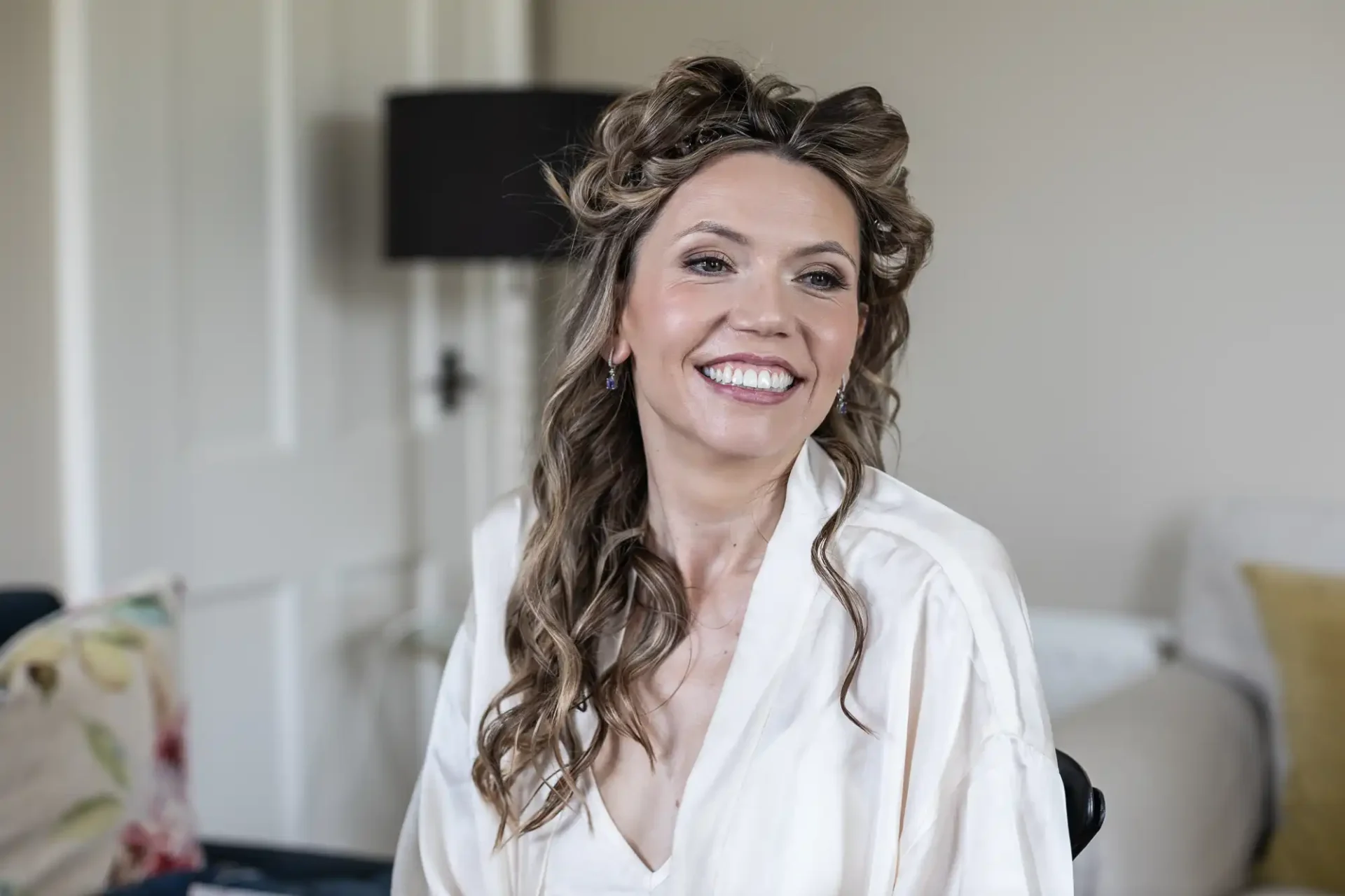 A woman with long, curly hair is smiling while sitting indoors. She is wearing a light-colored robe and appears relaxed.