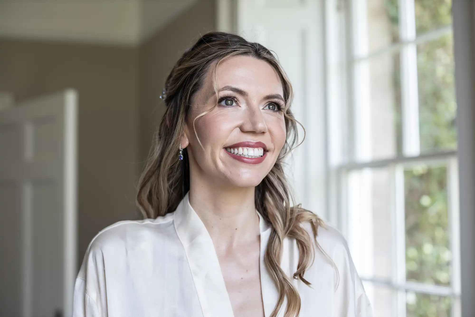A woman with long, wavy hair and light-colored robe is smiling and looking out a window. Natural light brightens the room.
