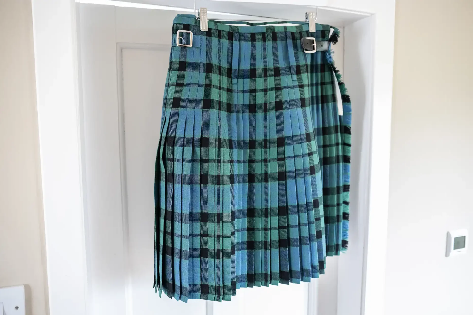 A blue and green tartan kilt with pleats and two silver buckles hangs on a white door.