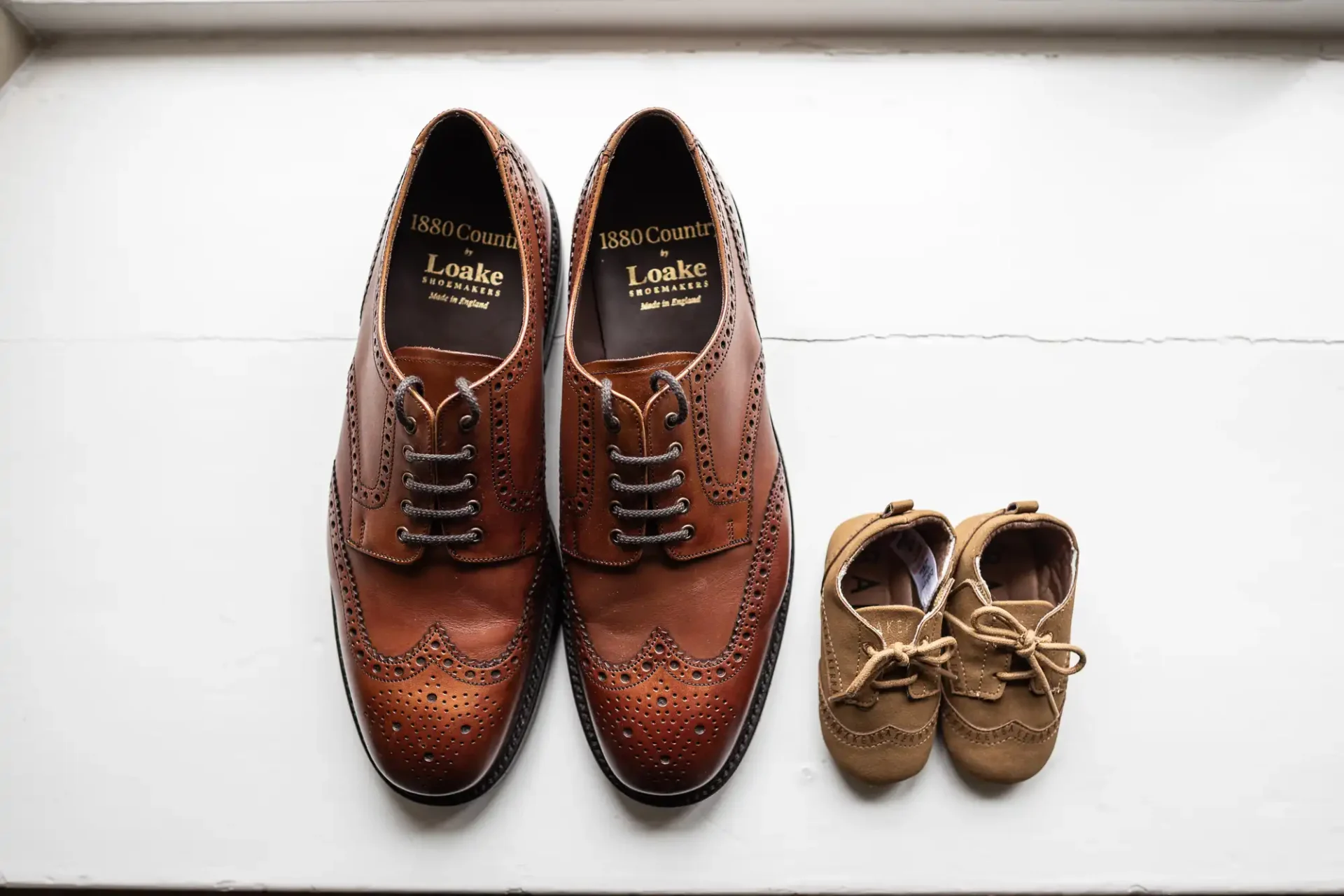 A pair of brown adult dress shoes is placed next to a smaller pair of tan baby moccasins on a white surface.