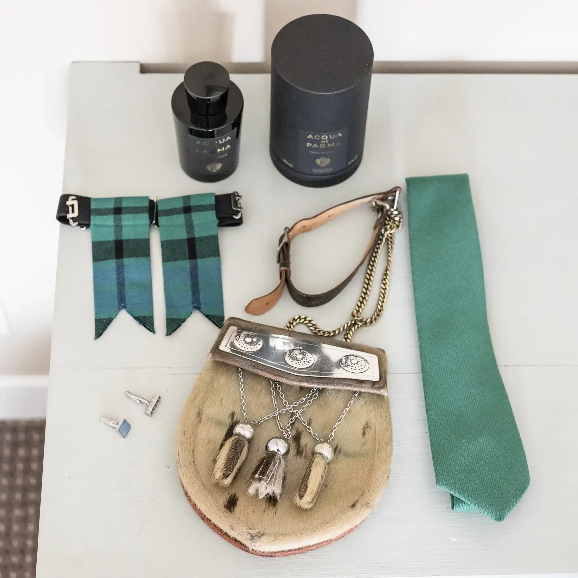 A green tie, plaid suspenders, a sporran, two bottles of cologne, cufflinks, and a leather strap are neatly arranged on a white surface.