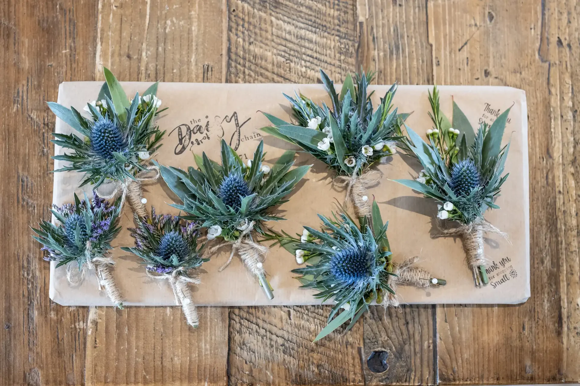Six small floral arrangements, each featuring a blue thistle surrounded by greenery, are neatly arranged on a brown paper sheet placed on a wooden surface.