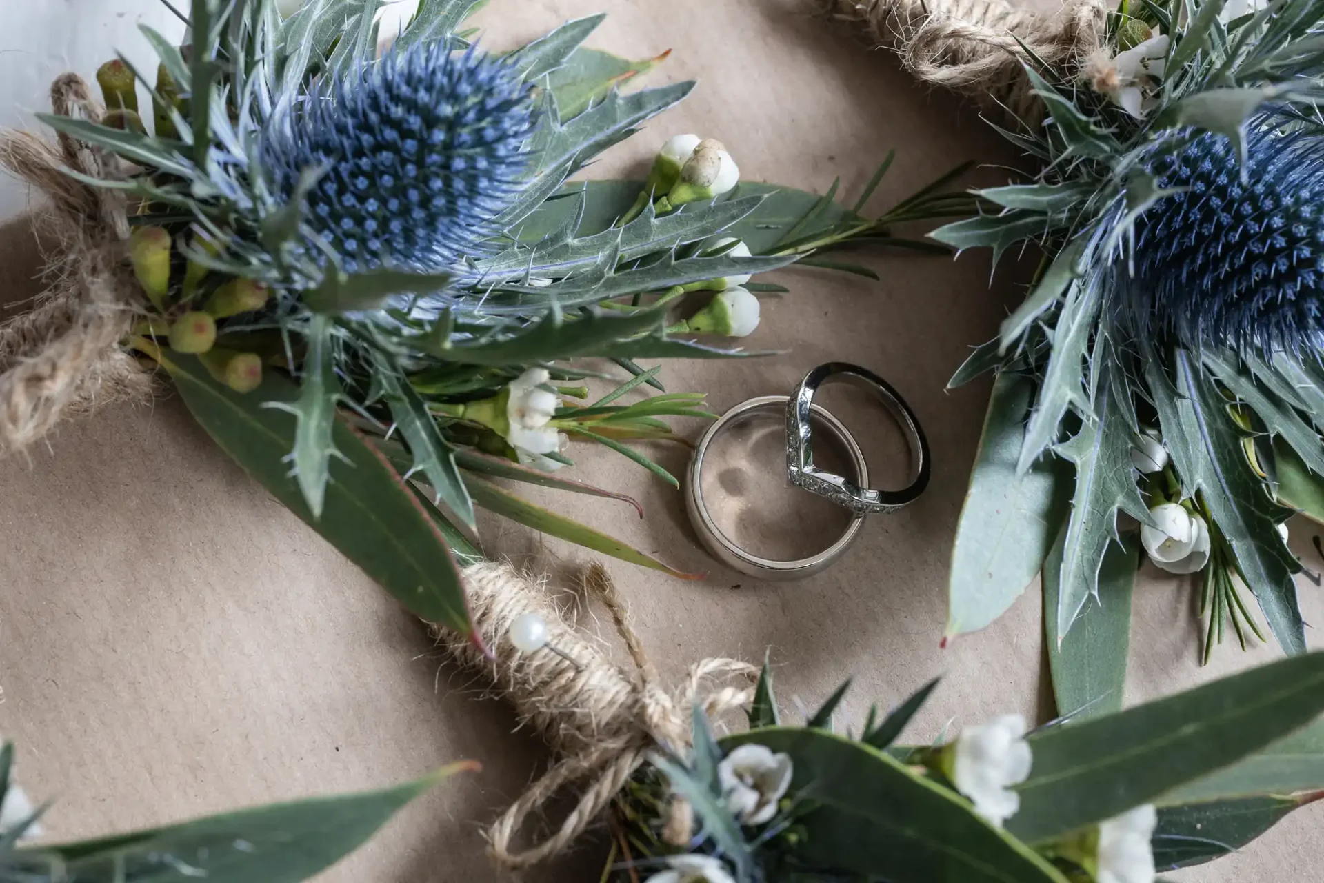 Two silver wedding rings are surrounded by a wreath of green foliage, blue thistle, and white flowers on a brown paper background.