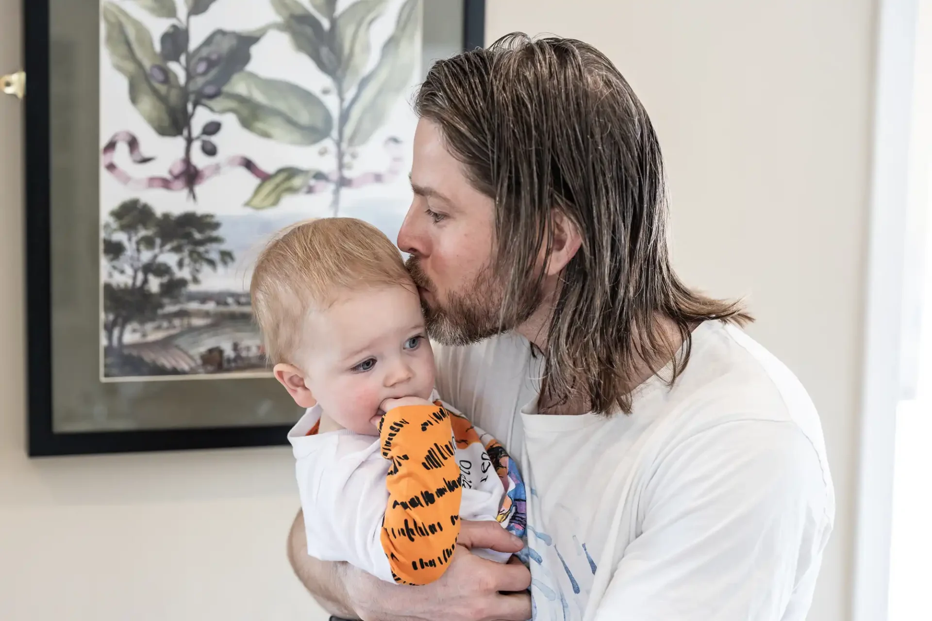 A man with long hair kisses a baby on the forehead while holding the baby in his arms. The baby is holding an orange object. A framed picture is visible in the background.