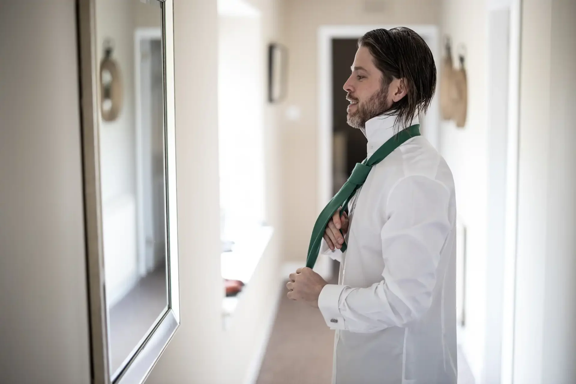 A man in a white shirt is tying a green tie while looking into a mirror in a hallway.