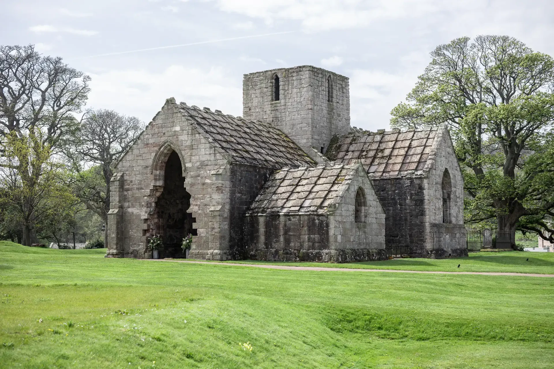 Old stone church surrounded by lush green grass and trees. The sky is cloudy. The building shows signs of age and wear.