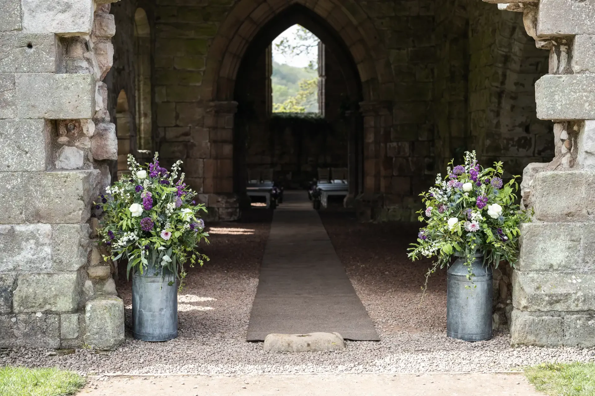 A stone archway leads to an interior space with two large floral arrangements in galvanized metal containers on either side of the entrance.