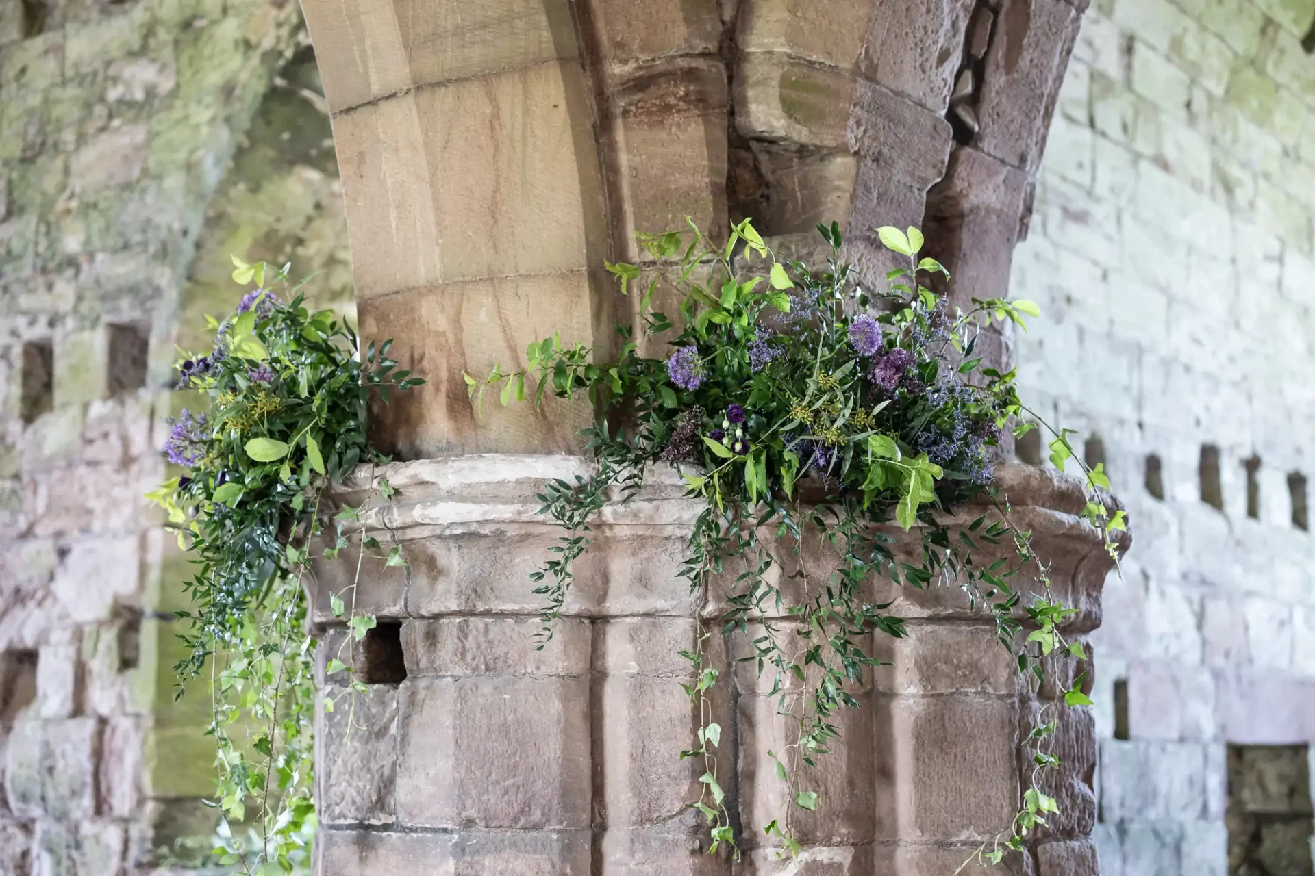Stone column decorated with green vines and purple flowers, situated in an old, rustic building with stone walls.