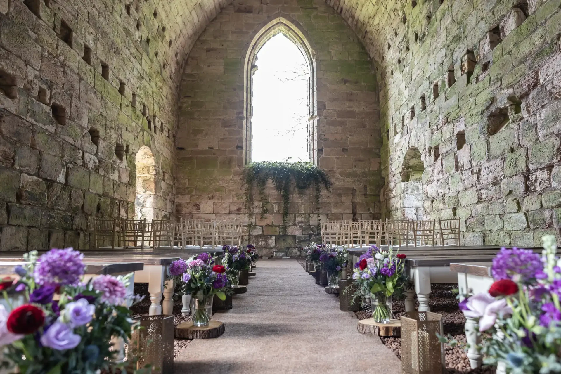 A rustic stone church interior with a high arched ceiling and arched window. The aisle is lined with bouquets of various flowers, and wooden chairs are arranged on either side.