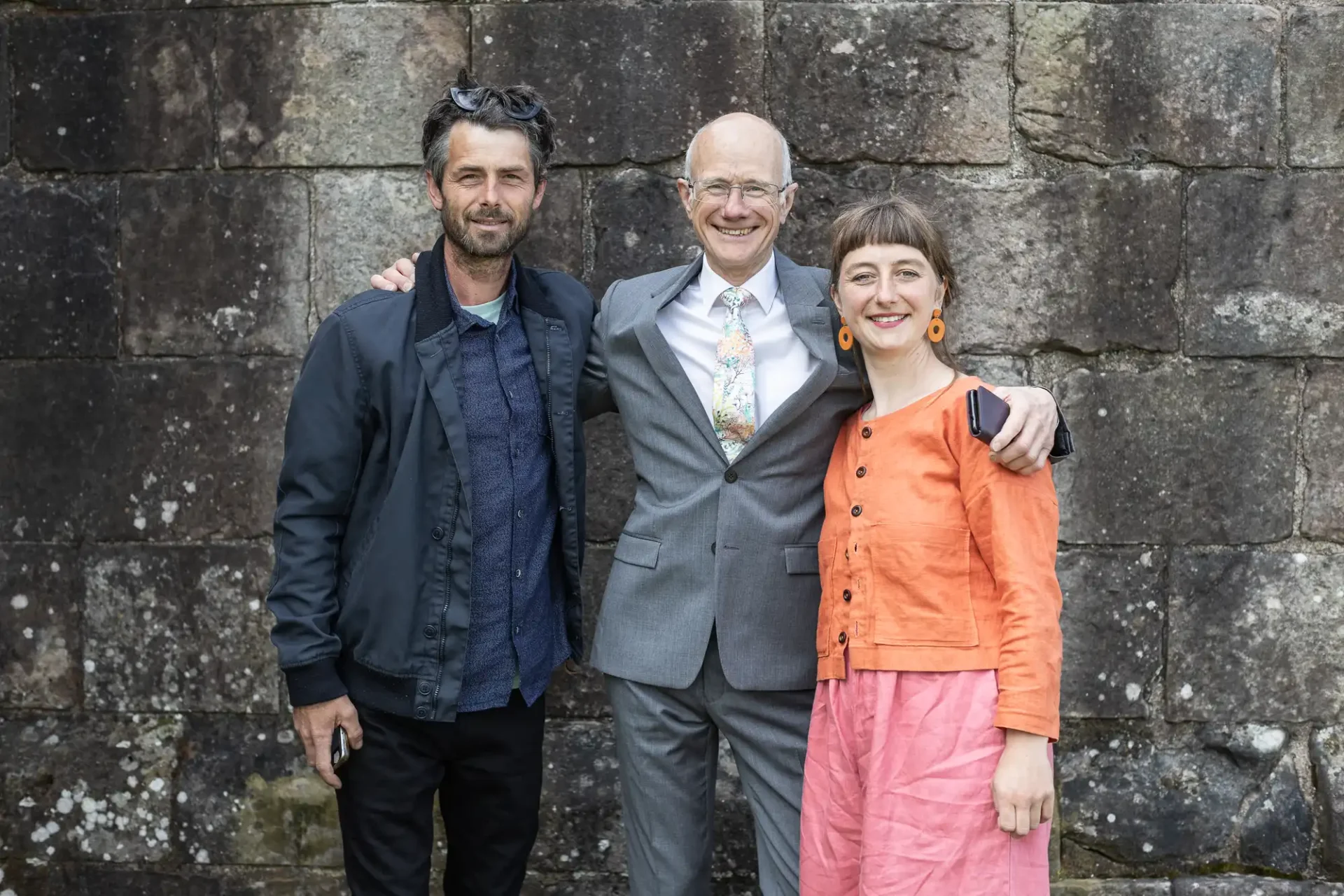 Three people standing in front of a stone wall; one man in a suit is in the center, with his arms around a man in a jacket on the left and a woman in an orange jacket and pink skirt on the right.
