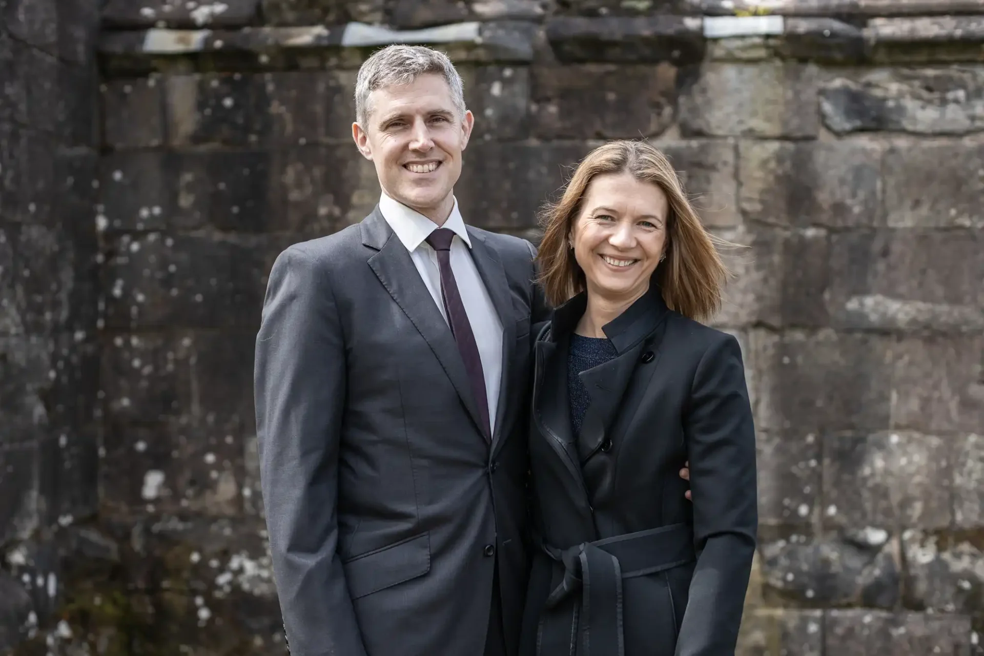 A man and a woman, both dressed in formal suits, stand arm in arm smiling in front of a stone wall.