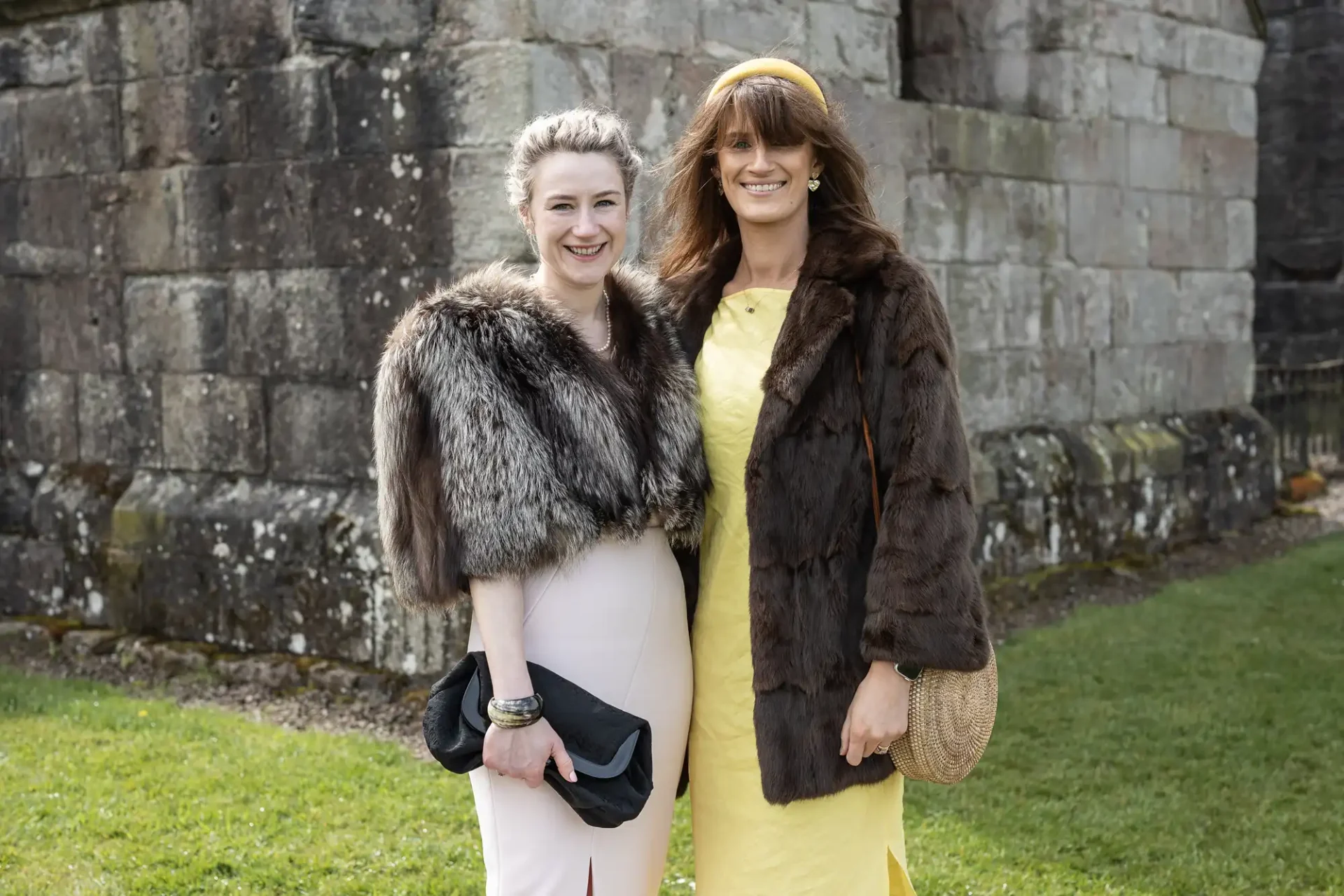 Two women stand outdoors in front of a stone wall, both wearing fur coats and smiling. One woman wears a pink dress and holds a clutch, the other wears a yellow dress and holds a woven bag.