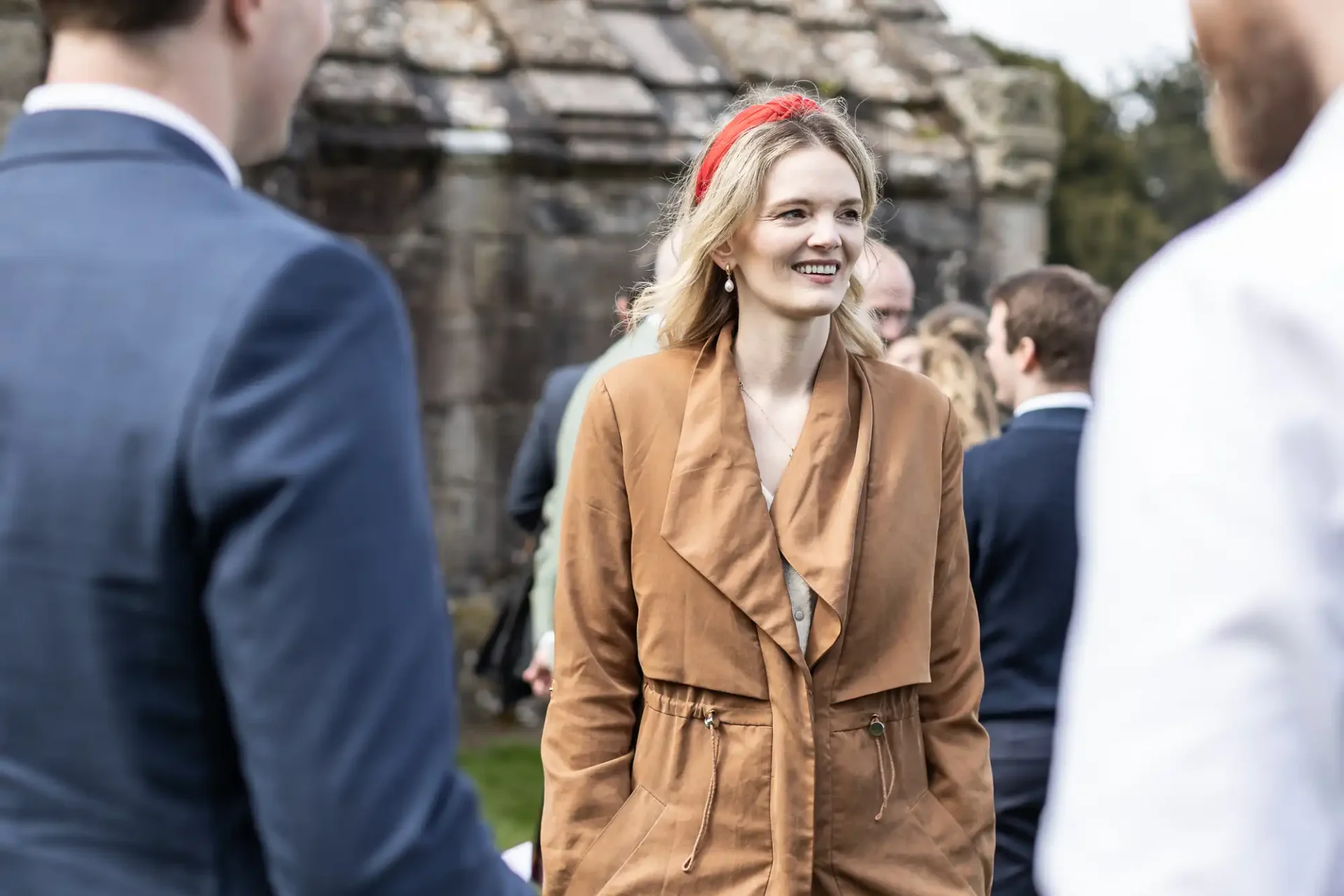 A woman wearing a brown coat and red headband smiles while standing among a group of people outdoors.