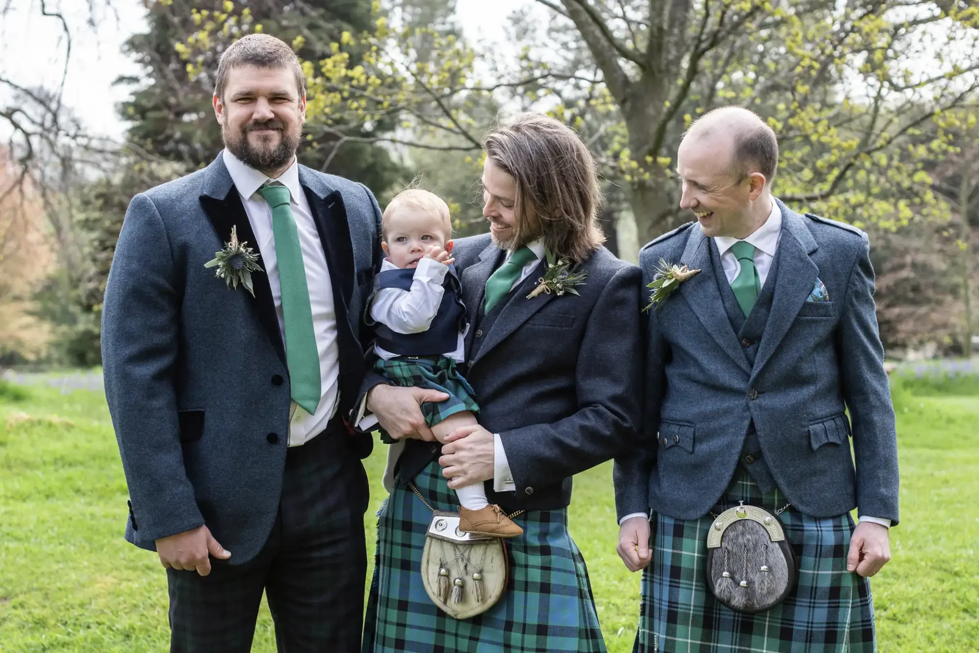 Three men and a baby, all dressed in kilts and matching green ties, standing outside on grass with trees in the background. The baby is being held by the man in the center.
