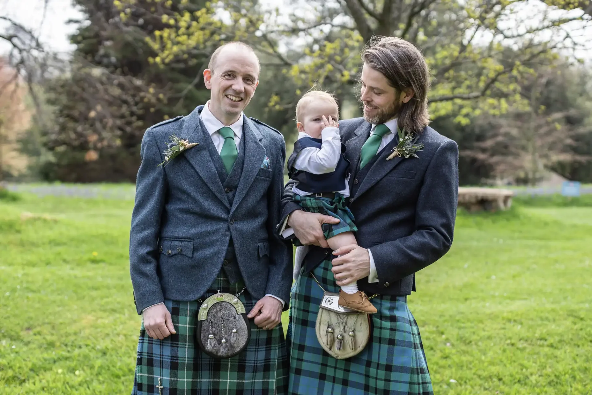Two men dressed in traditional Scottish kilts stand outdoors, one holding a baby who is also in a kilt. Trees and greenery are visible in the background.
