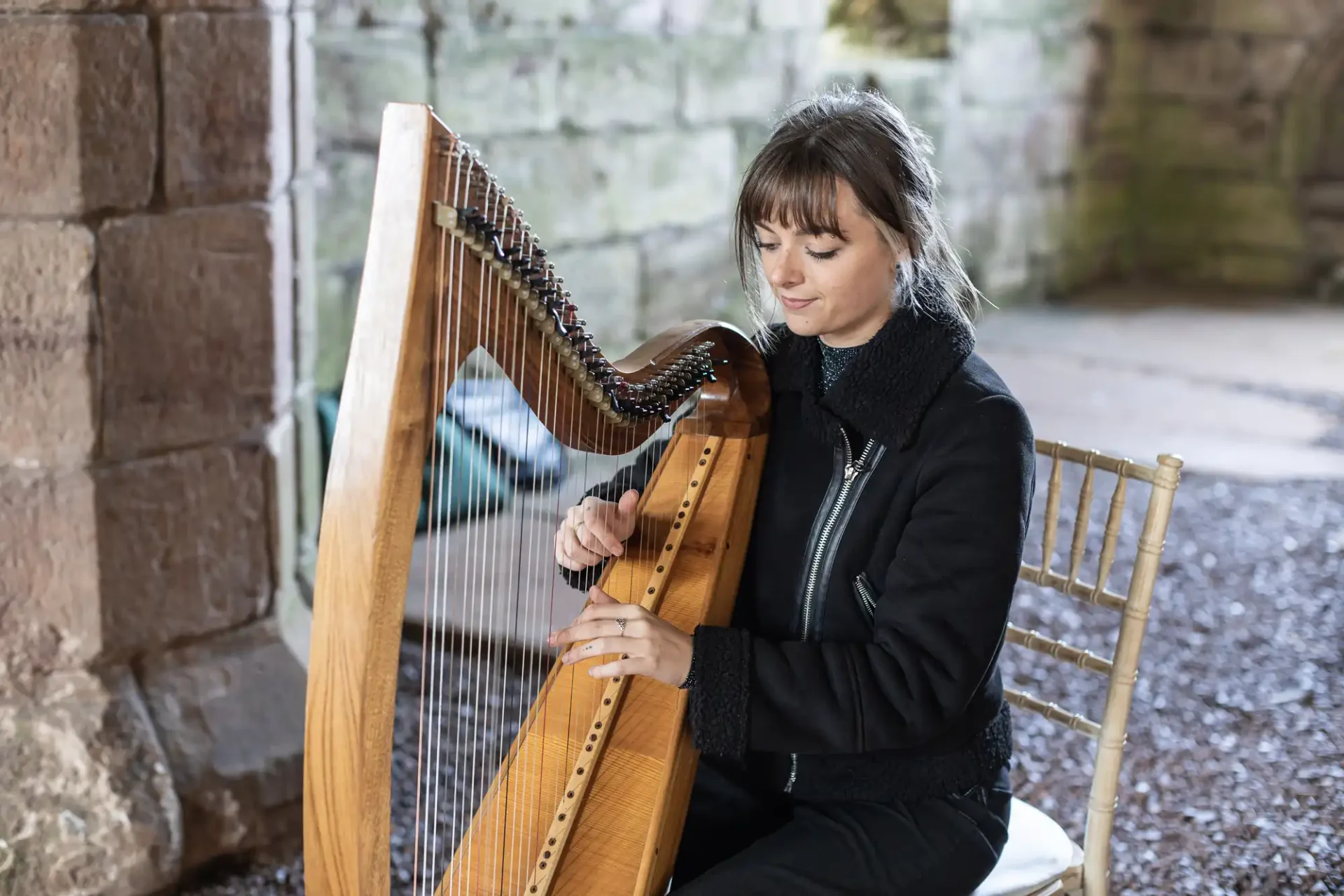 A woman sits on a wooden chair playing a harp, with stone walls in the background.