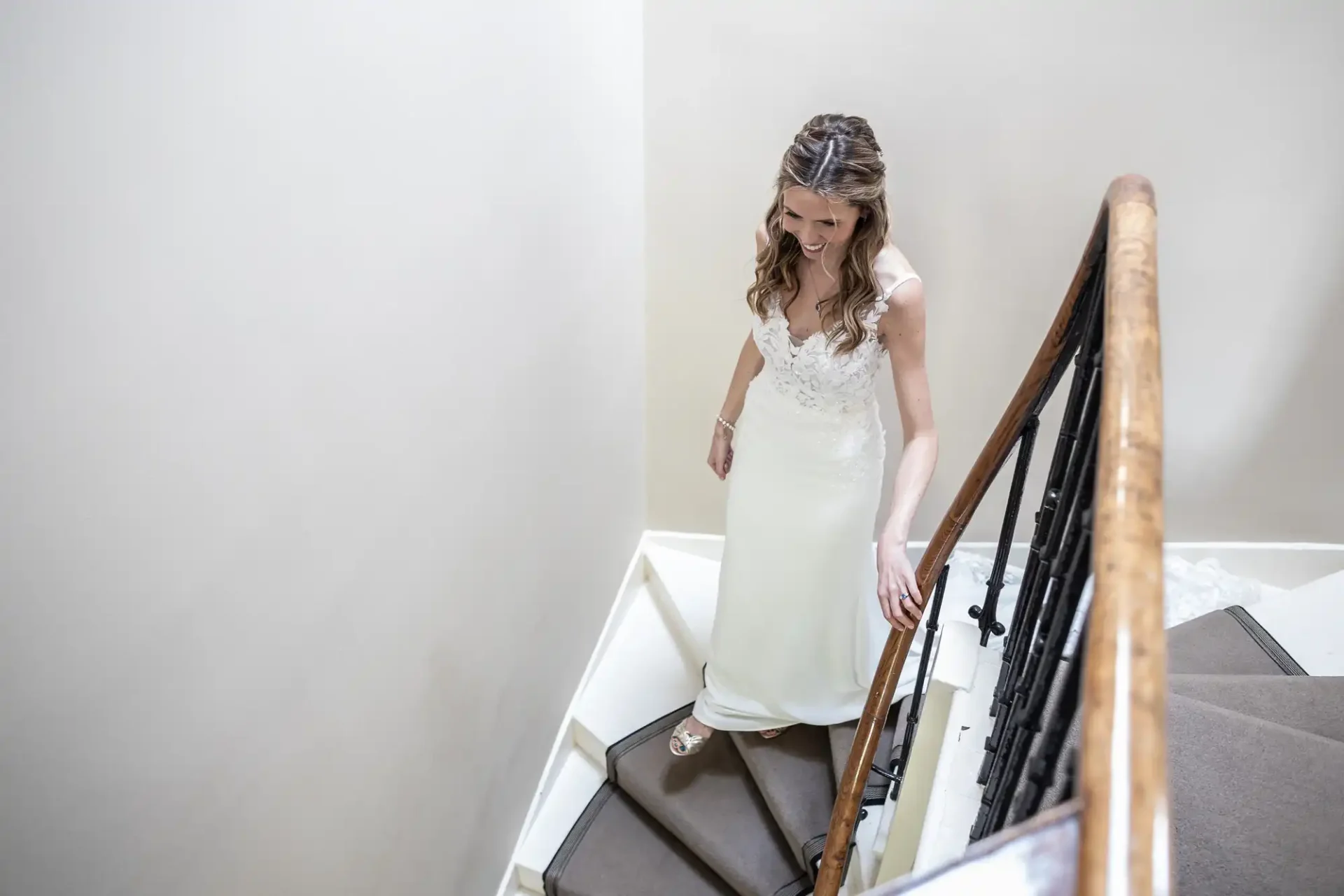 A woman in a white wedding dress ascends a staircase, holding the handrail and smiling.