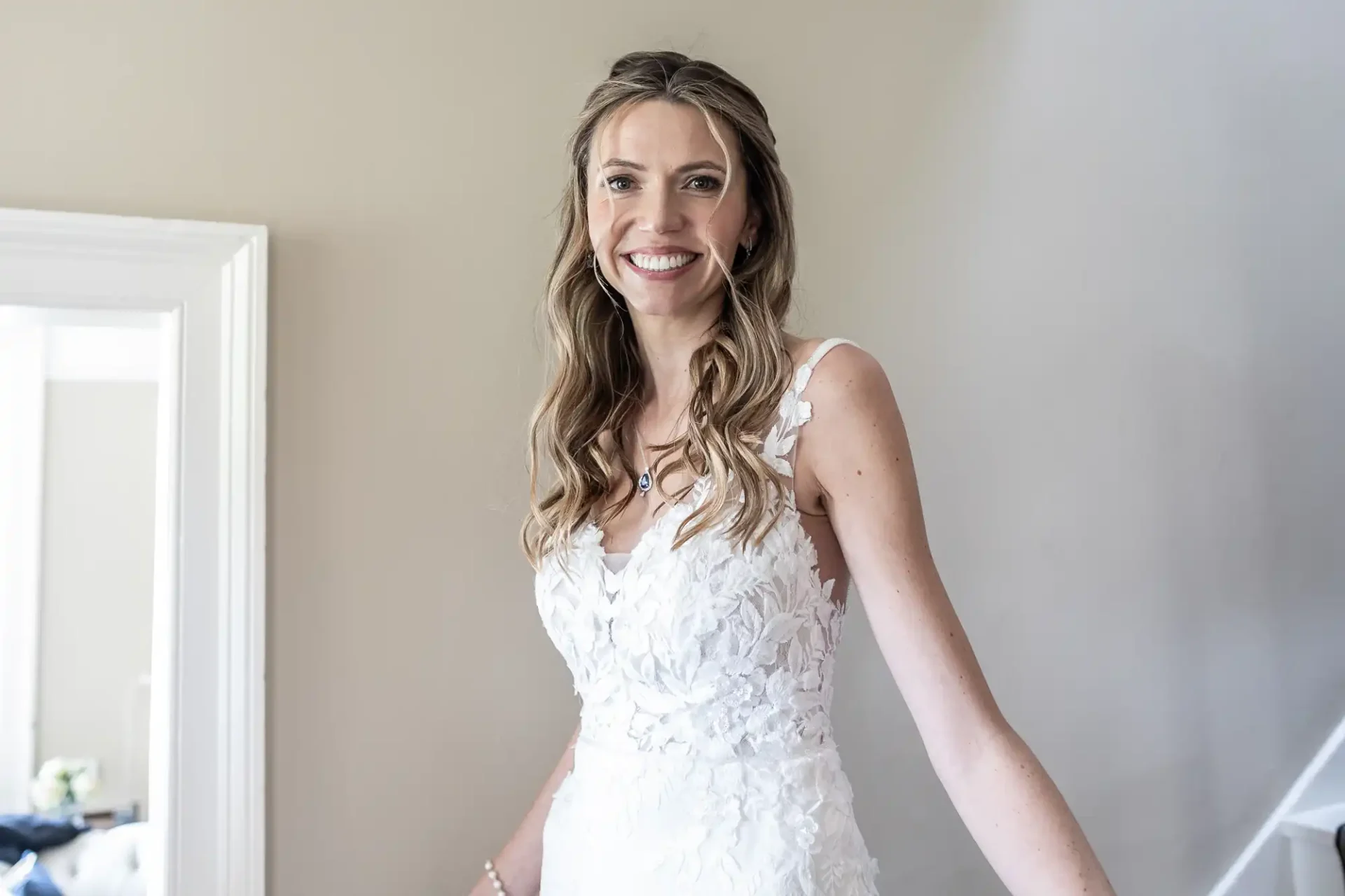 A woman in a white lace wedding dress stands indoors, smiling at the camera.