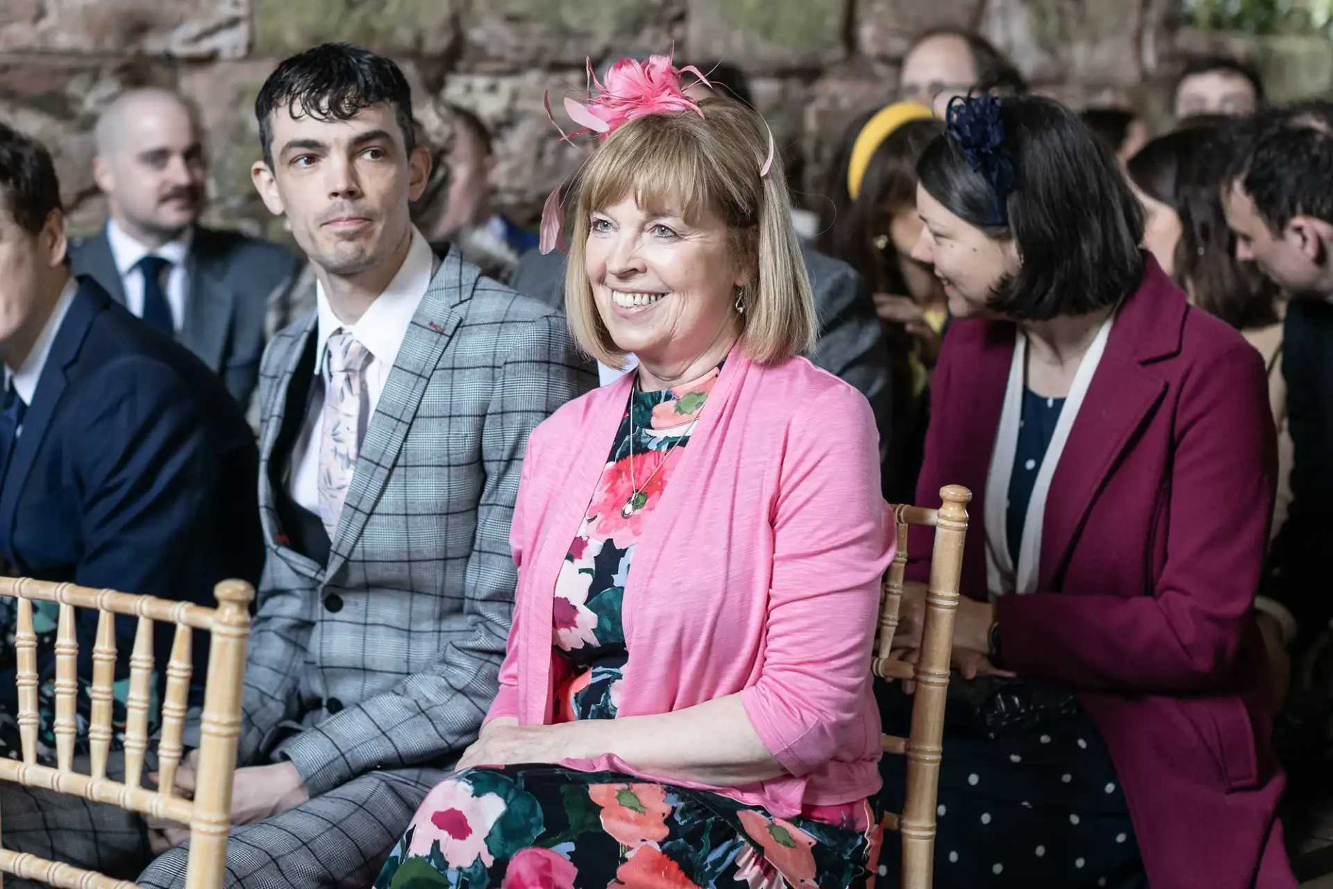 A group of people sitting on wooden chairs at an event. A woman in the foreground wears a floral dress and pink cardigan, with a matching pink fascinator, smiling.