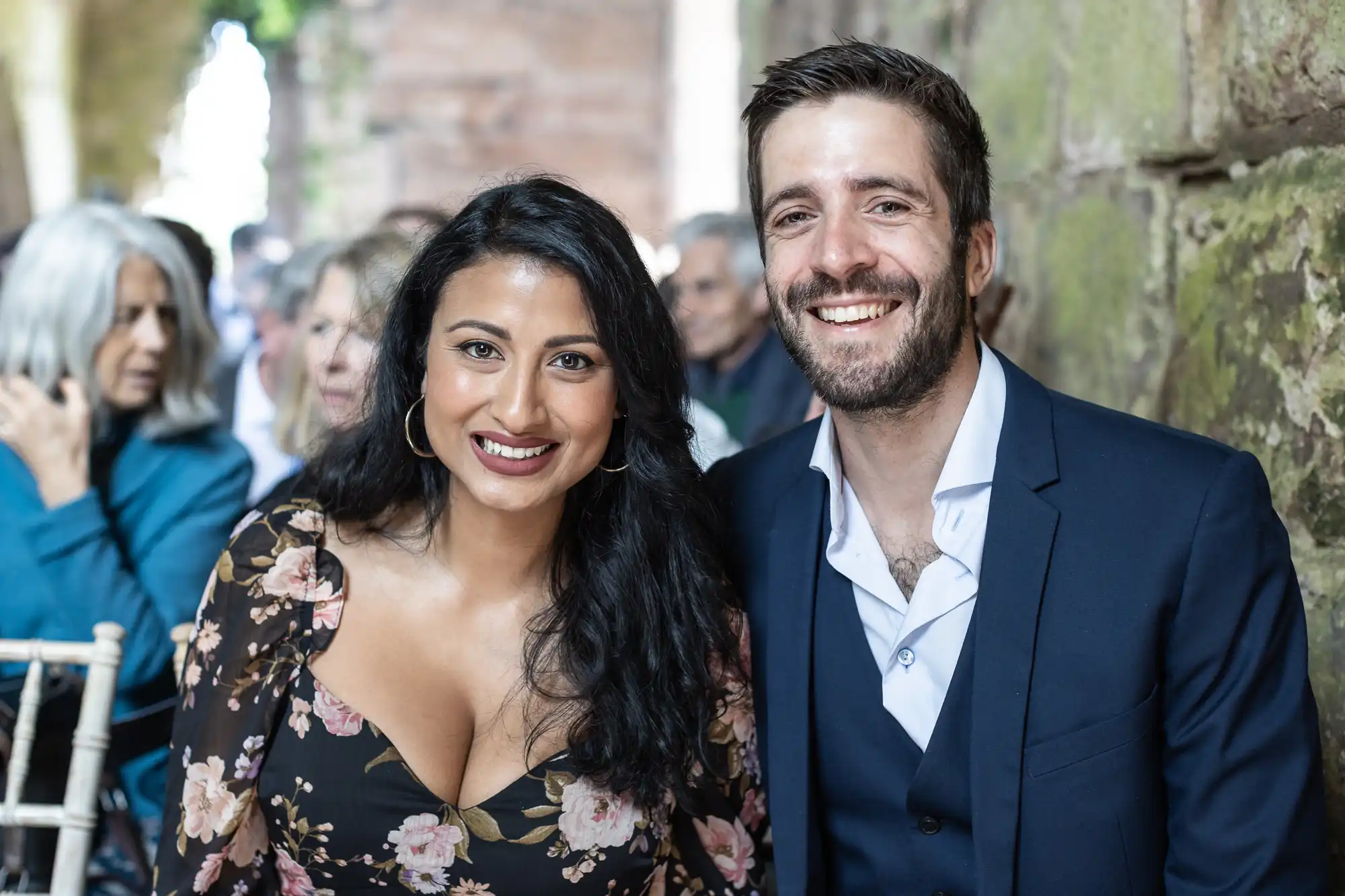 A woman in a floral dress and a man in a dark suit smile at the camera, standing close together in an outdoor setting with stone walls and a crowd in the background.
