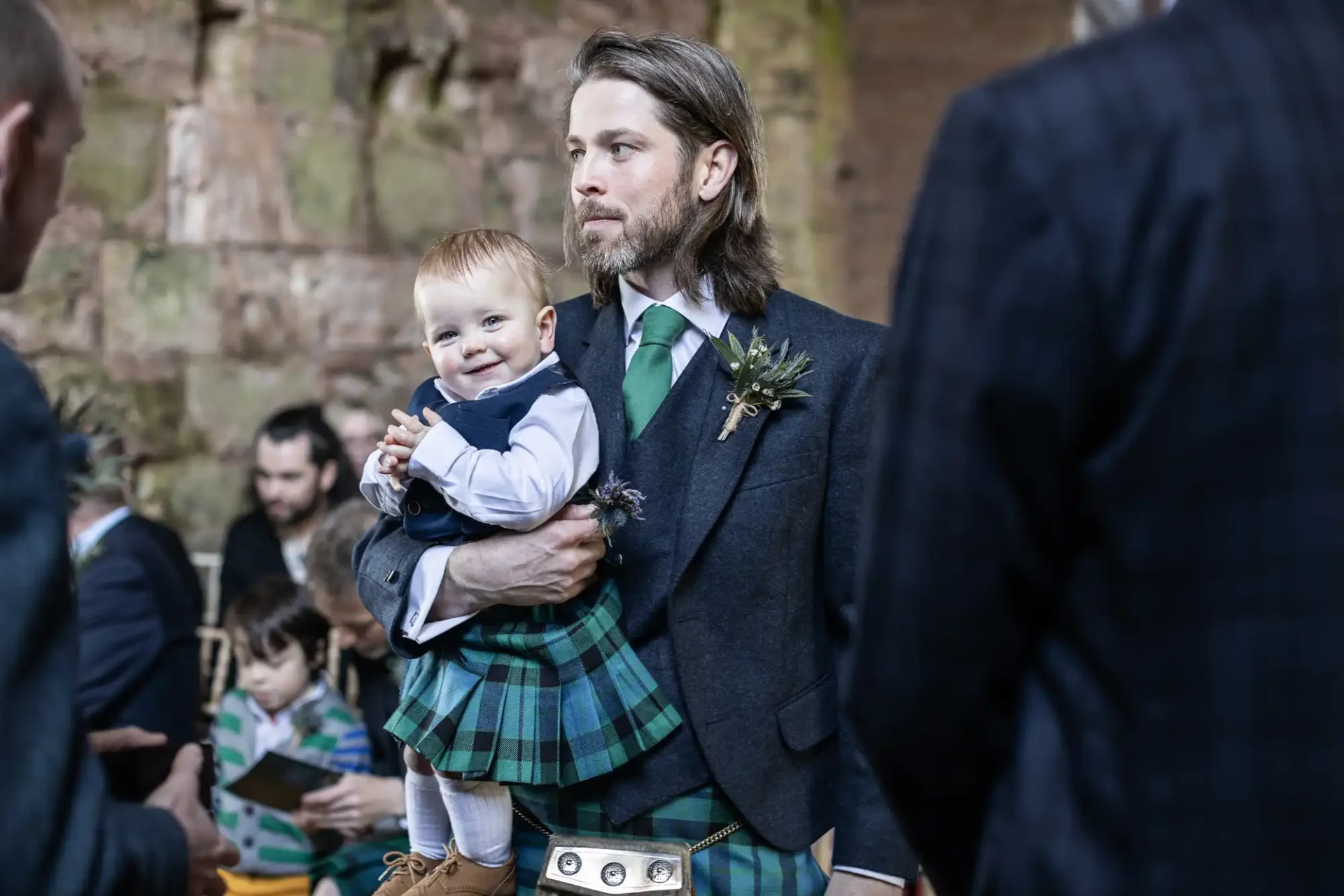 A man in a traditional kilt holds a smiling baby also dressed in a similar outfit. They are indoors with other people in the background.