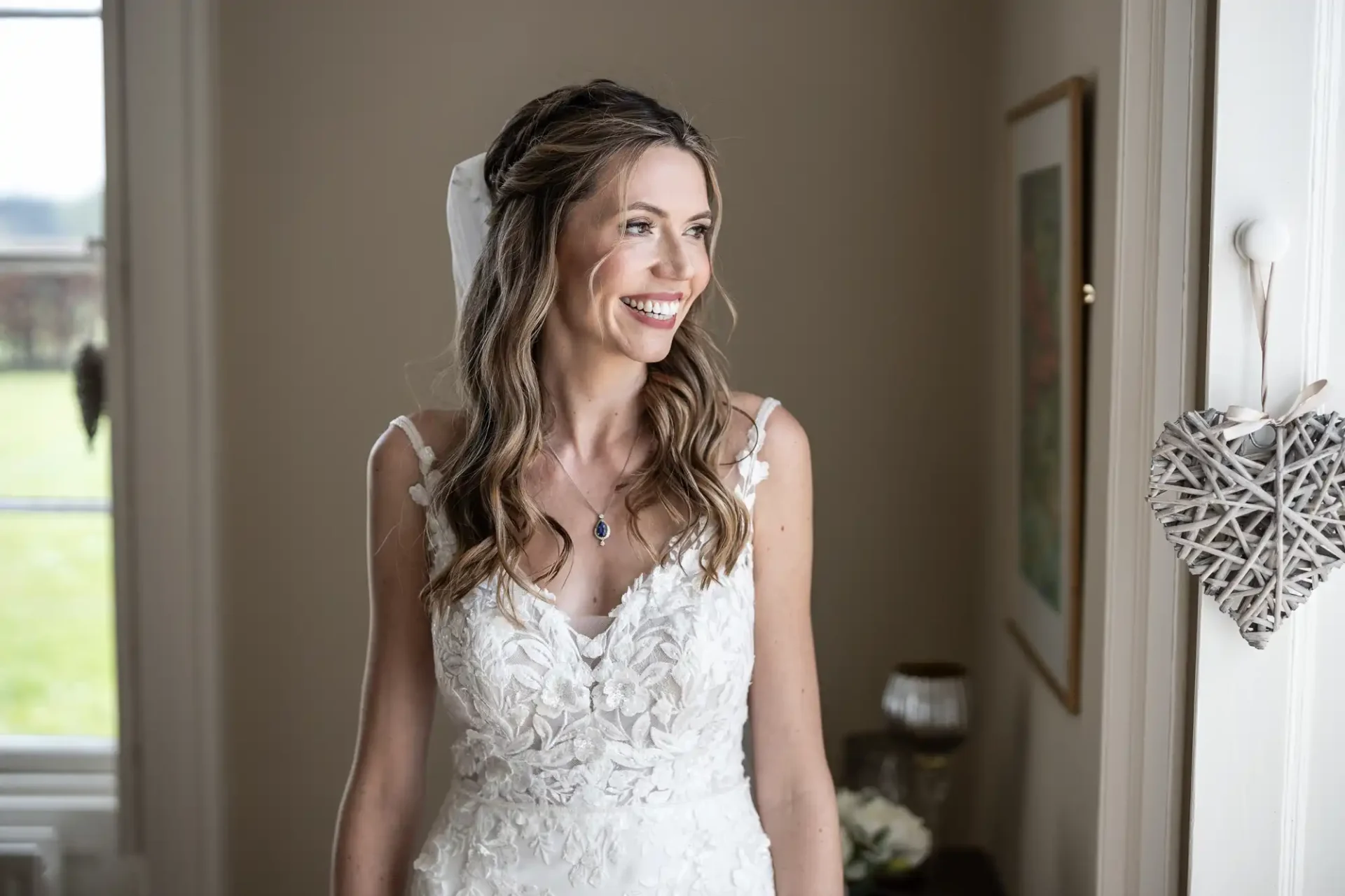 A bride with long wavy hair smiles while standing in a room decorated with a heart-shaped ornament and white flowers. She is wearing a sleeveless white lace wedding dress.