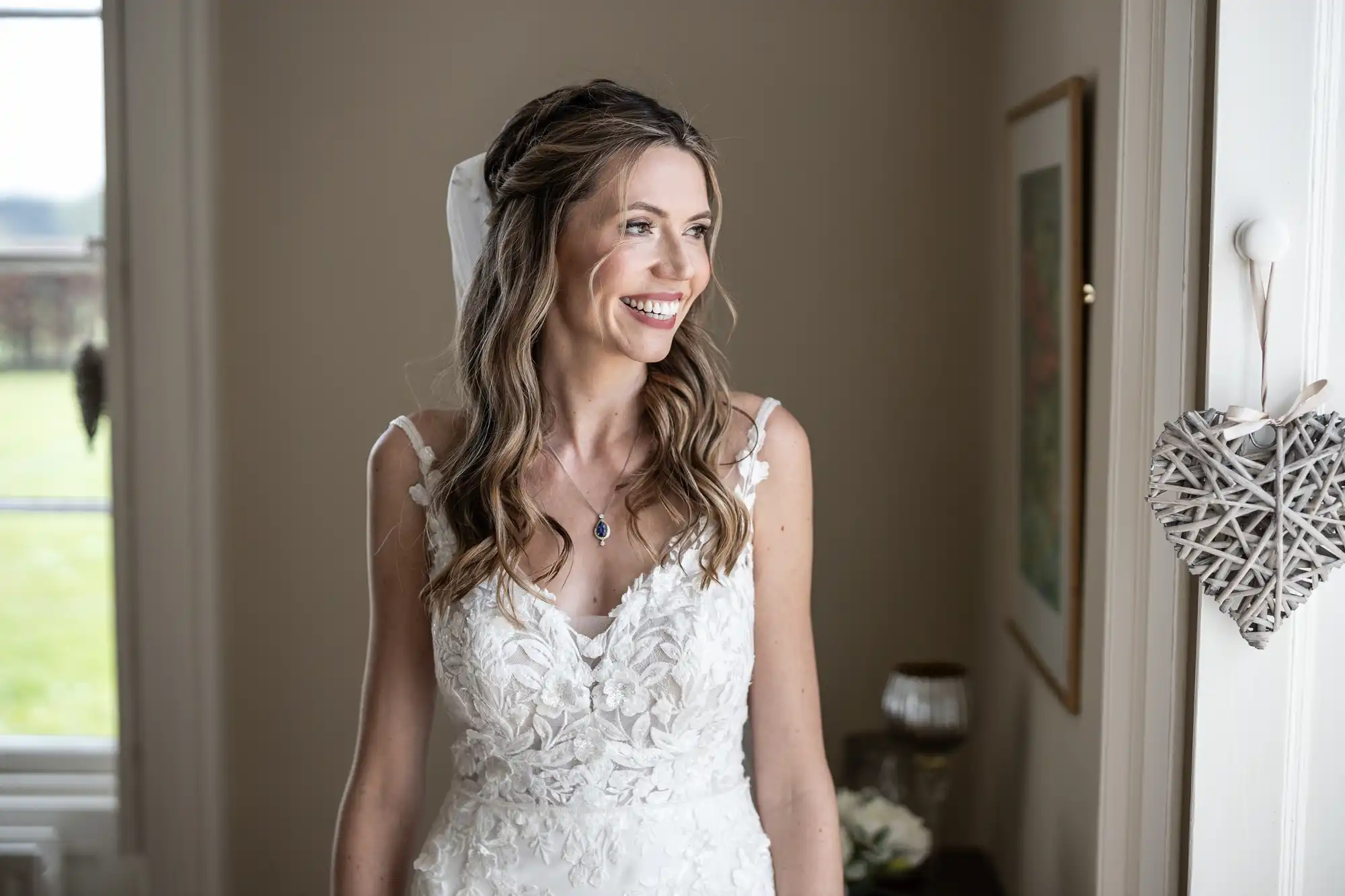 Wedding photos gallery featuring a bride with long wavy hair smiles while standing in a room decorated with a heart-shaped ornament and white flowers. She is wearing a sleeveless white lace wedding dress.