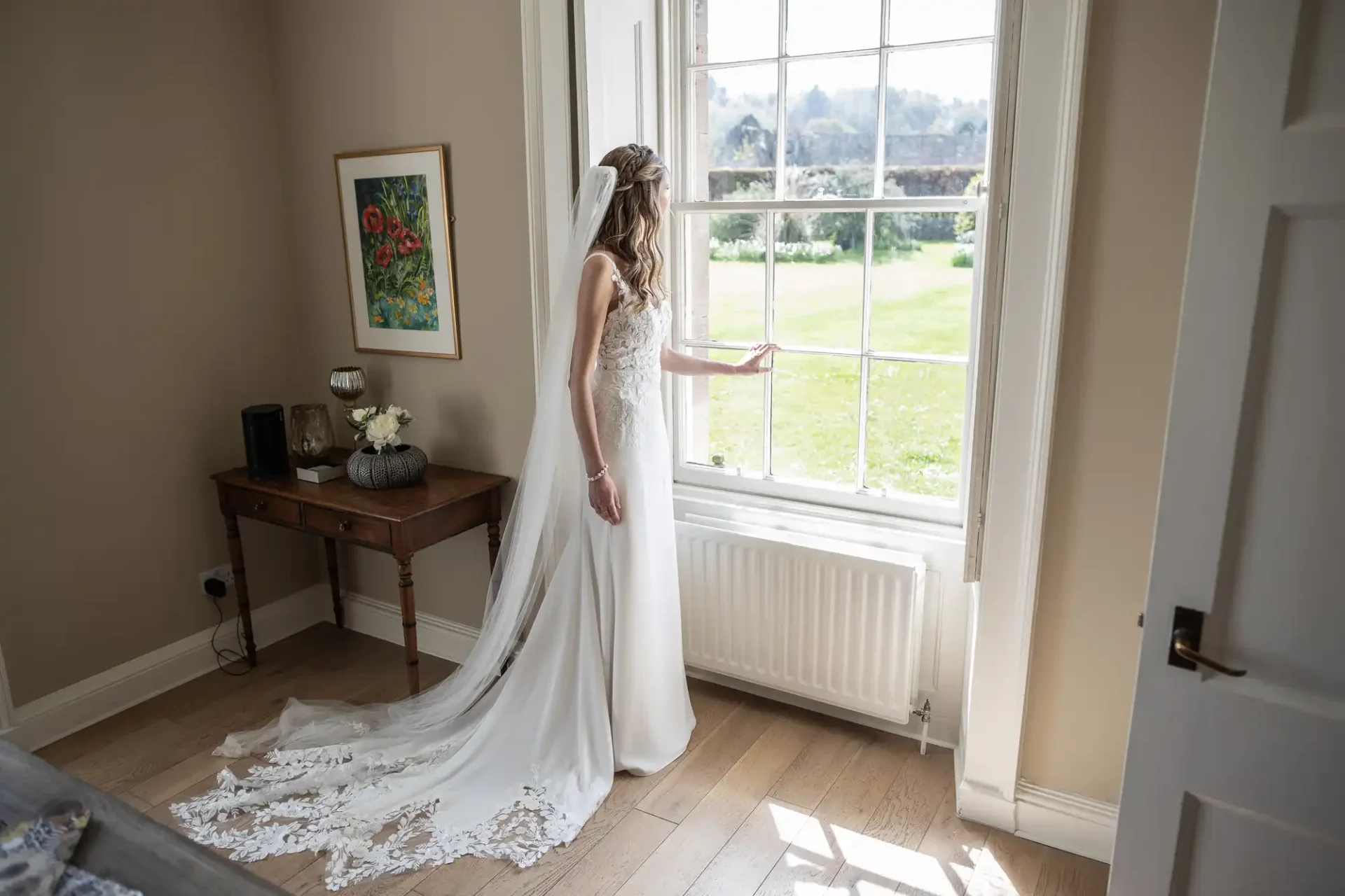 A bride in a white gown stands by a tall window, gazing outside, with her hand resting on the window frame. A small table with flowers and artwork decorates the room.
