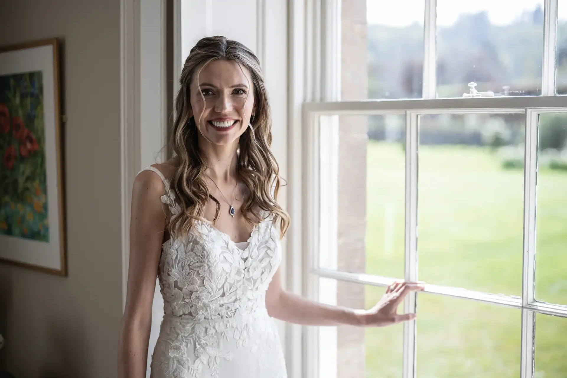 A woman in a white bridal gown stands beside a large window, smiling. She has long wavy hair and wears a pendant necklace. The background shows a green outdoor scene through the window.