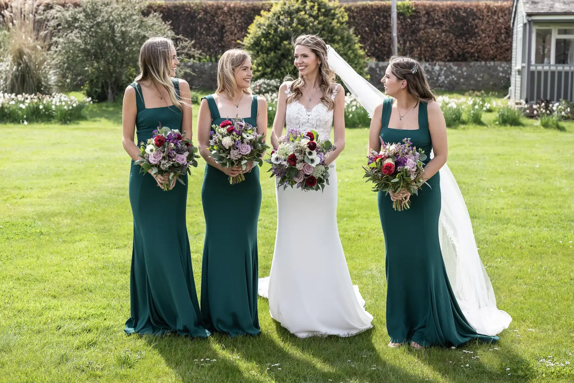 A bride in a white dress stands outside on grass with three bridesmaids in matching green dresses, all holding bouquets.