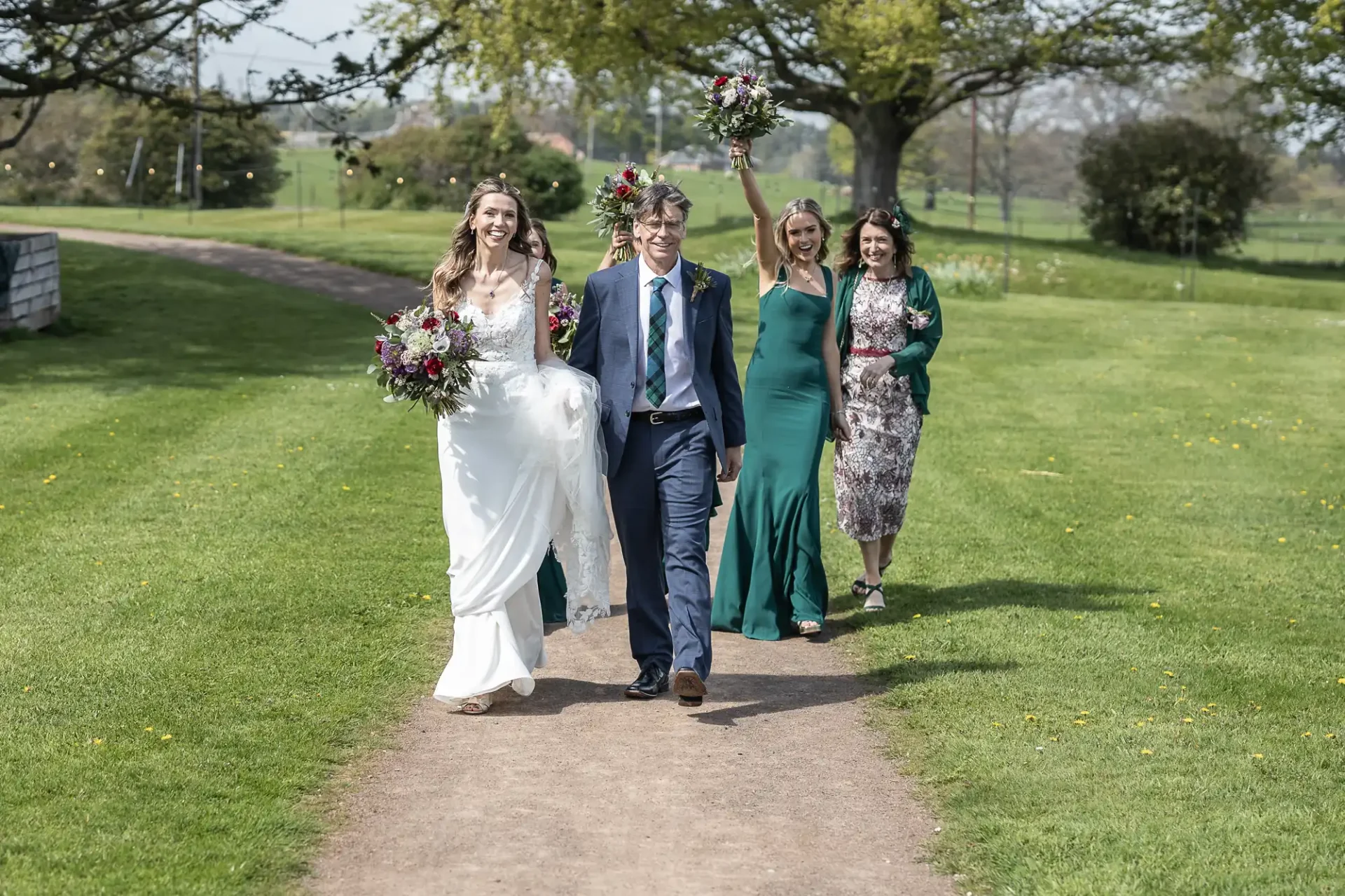 A newlywed couple walks outdoors, smiling, with three guests behind them. The bride holds a bouquet and wears a white dress; the groom is in a suit. The three guests wear green dresses and a patterned dress.