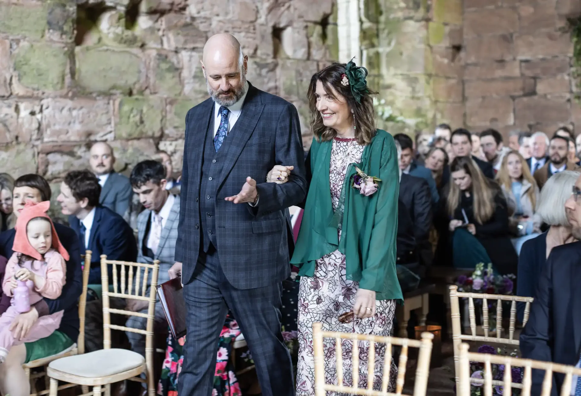 A man in a dark plaid suit and a woman in a green jacket walk down an aisle at an indoor gathering. They are surrounded by seated attendees facing them. The background features a stone wall.