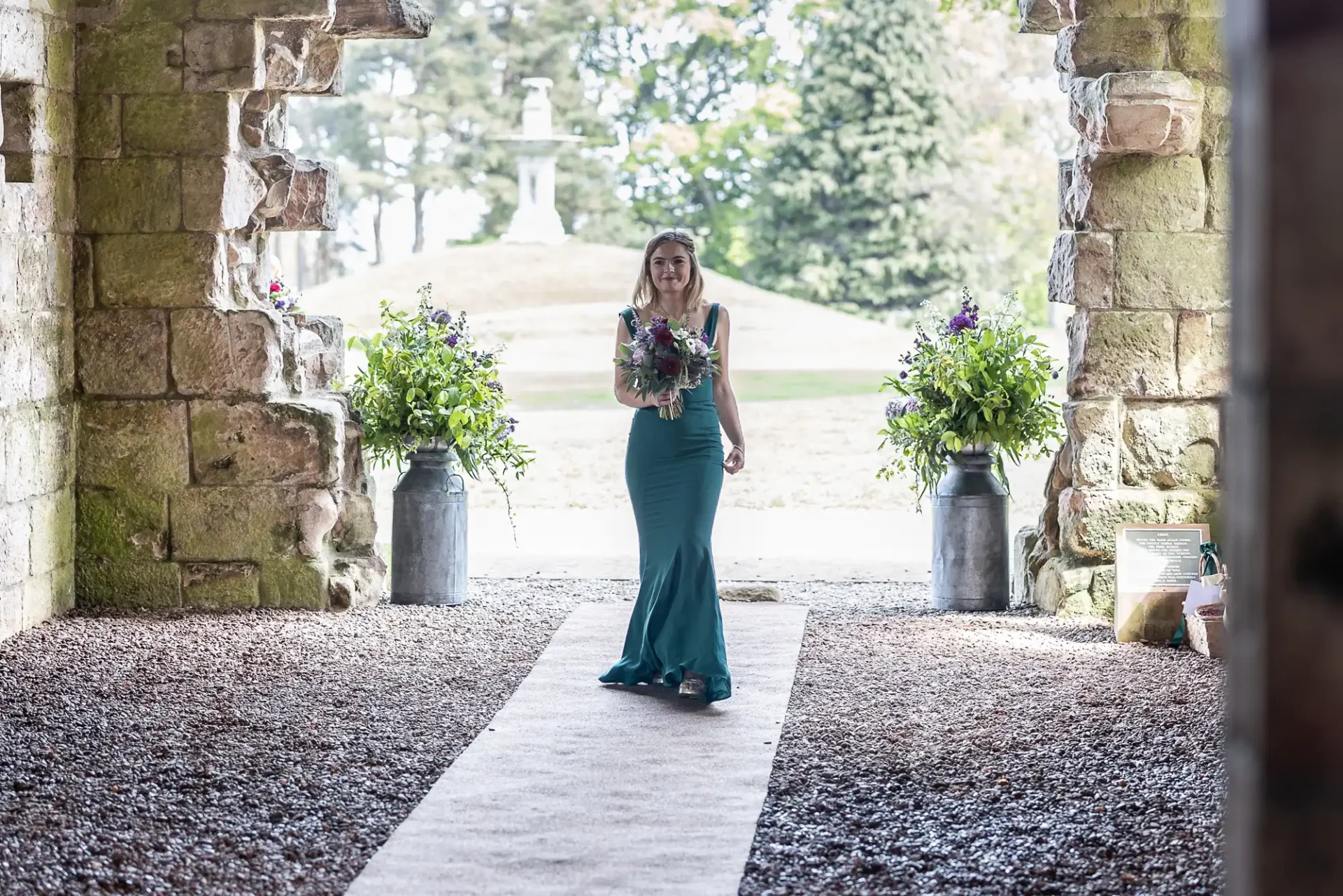 A woman in a teal dress walks down an aisle, holding a bouquet, surrounded by stone walls and large flower arrangements. Outdoor background visible through archway.