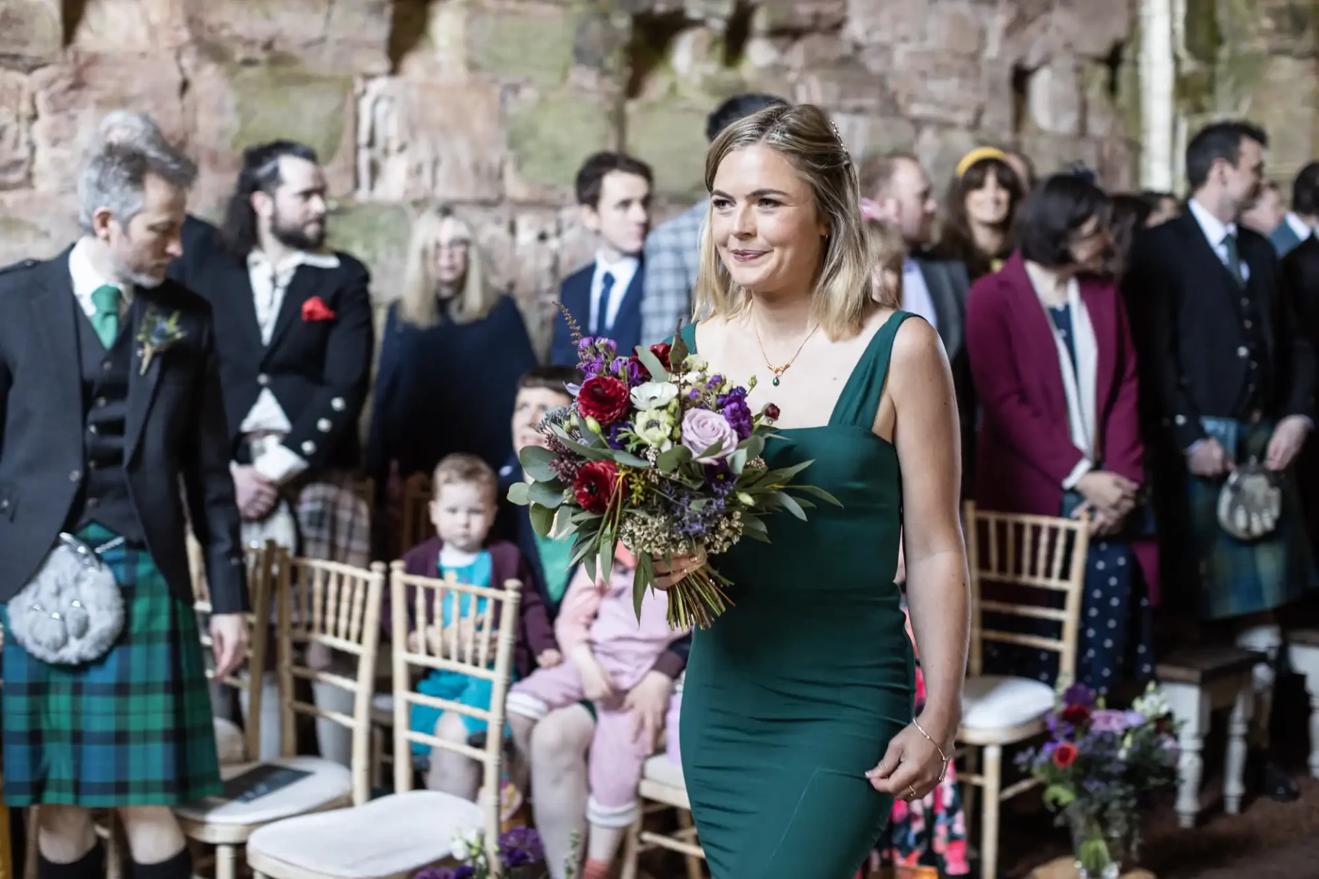 A woman in a green dress walks down an aisle, holding a bouquet of flowers. Guests, including men in kilts and children, are seated on both sides in a rustic setting with stone walls.