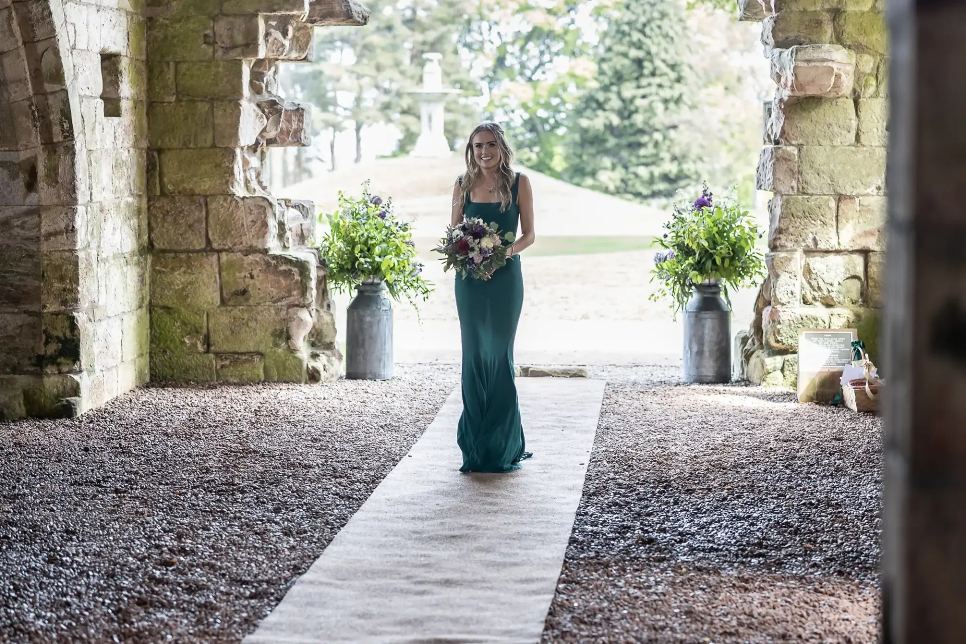 A woman in a long teal dress walks down an outdoor aisle, holding a bouquet, with stone archways and greenery in the background.