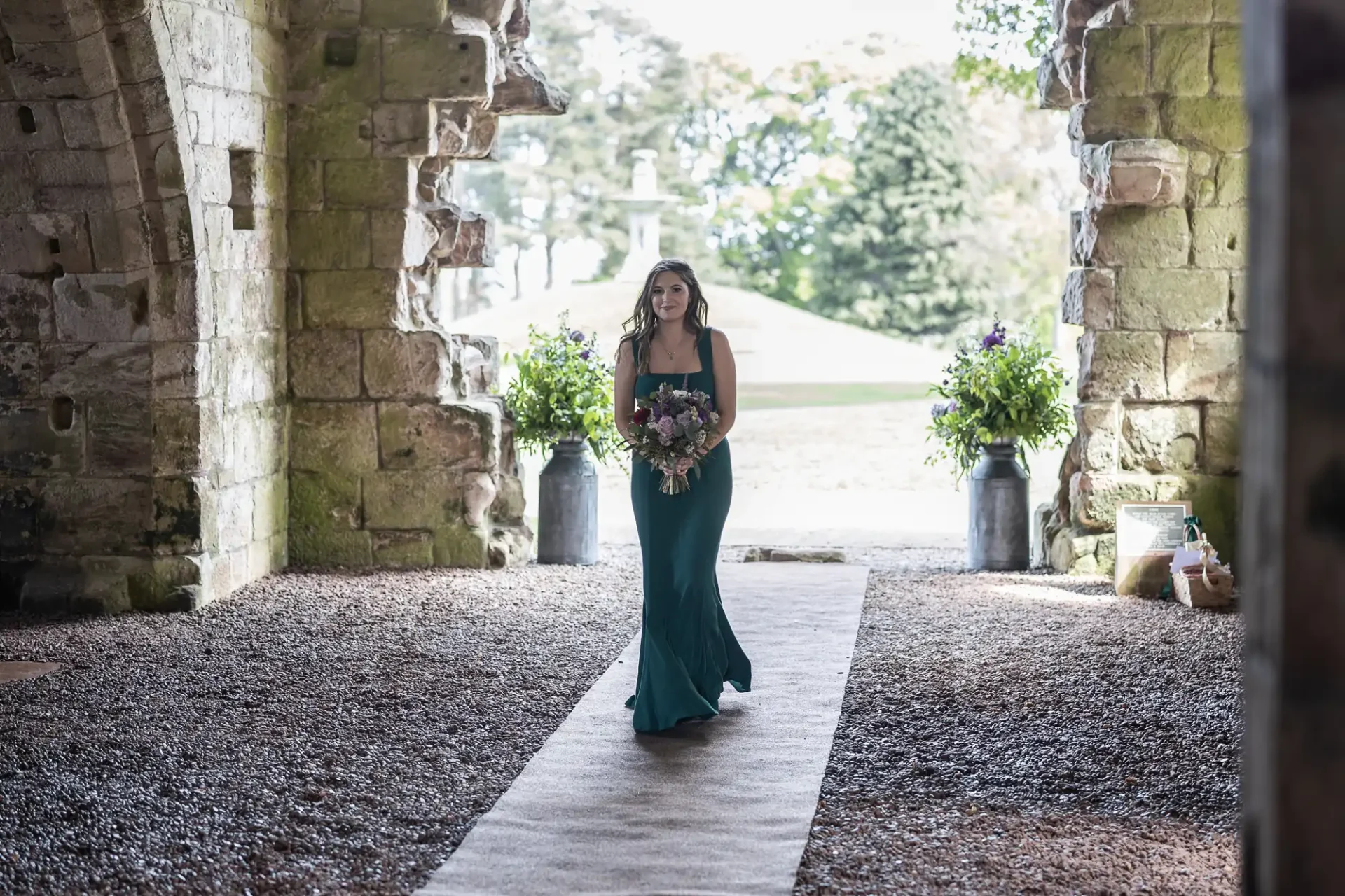 A woman in a green dress walks down a carpeted aisle inside a stone archway, holding a bouquet of flowers.