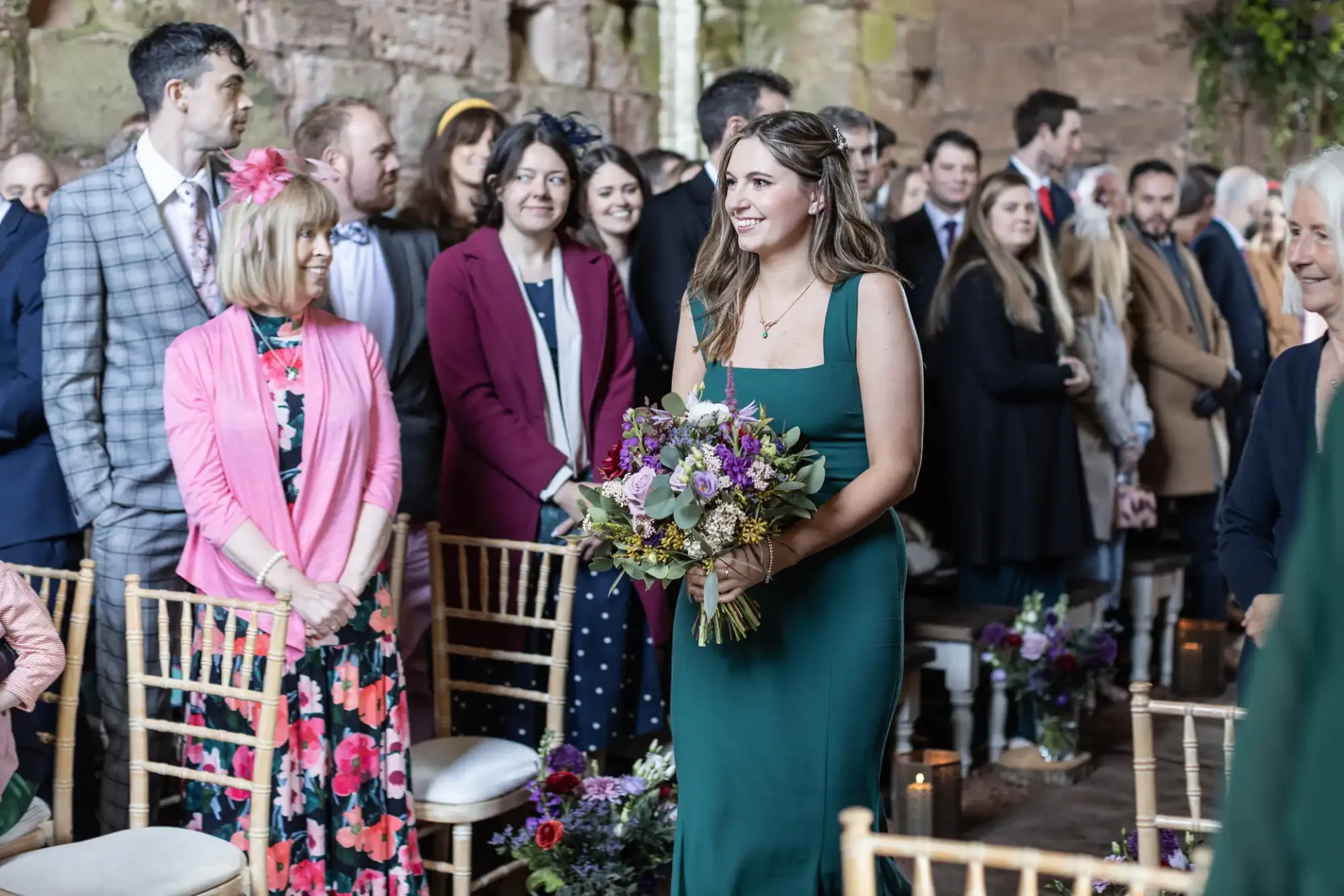 A woman in a green dress holds a bouquet while walking down an aisle at a wedding ceremony, with guests standing and seated nearby.