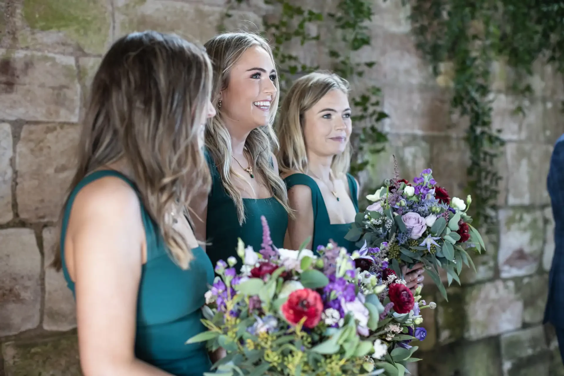 Three women in green dresses holding colorful flower bouquets stand in front of a stone wall adorned with greenery.