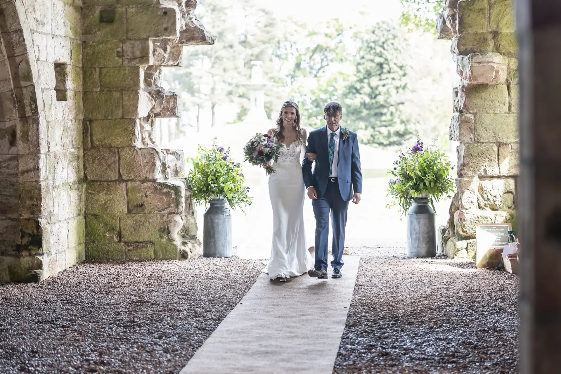 A bride and an older man walk down an outdoor aisle. The bride wears a white dress and holds a bouquet. They are framed by stone archways and greenery.