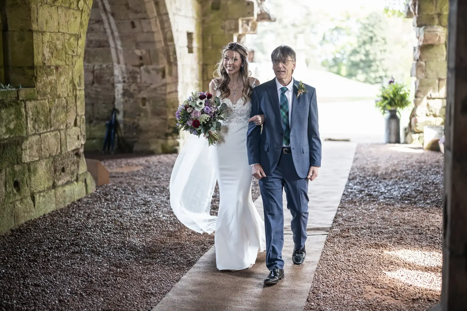 A bride holding a bouquet and an older man walk down an outdoor aisle under a stone archway. The bride wears a white gown and veil; the man is dressed in a blue suit and tie.