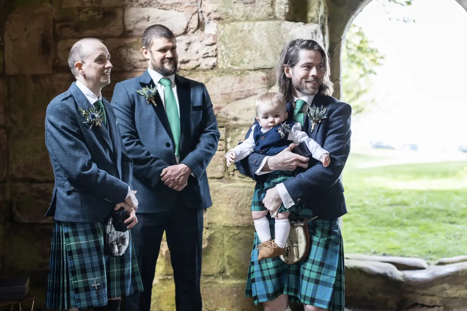 Three men wearing kilts stand near a stone wall. One man holds a baby, also dressed in a kilt. They appear to be outdoors near an archway with greenery visible beyond.