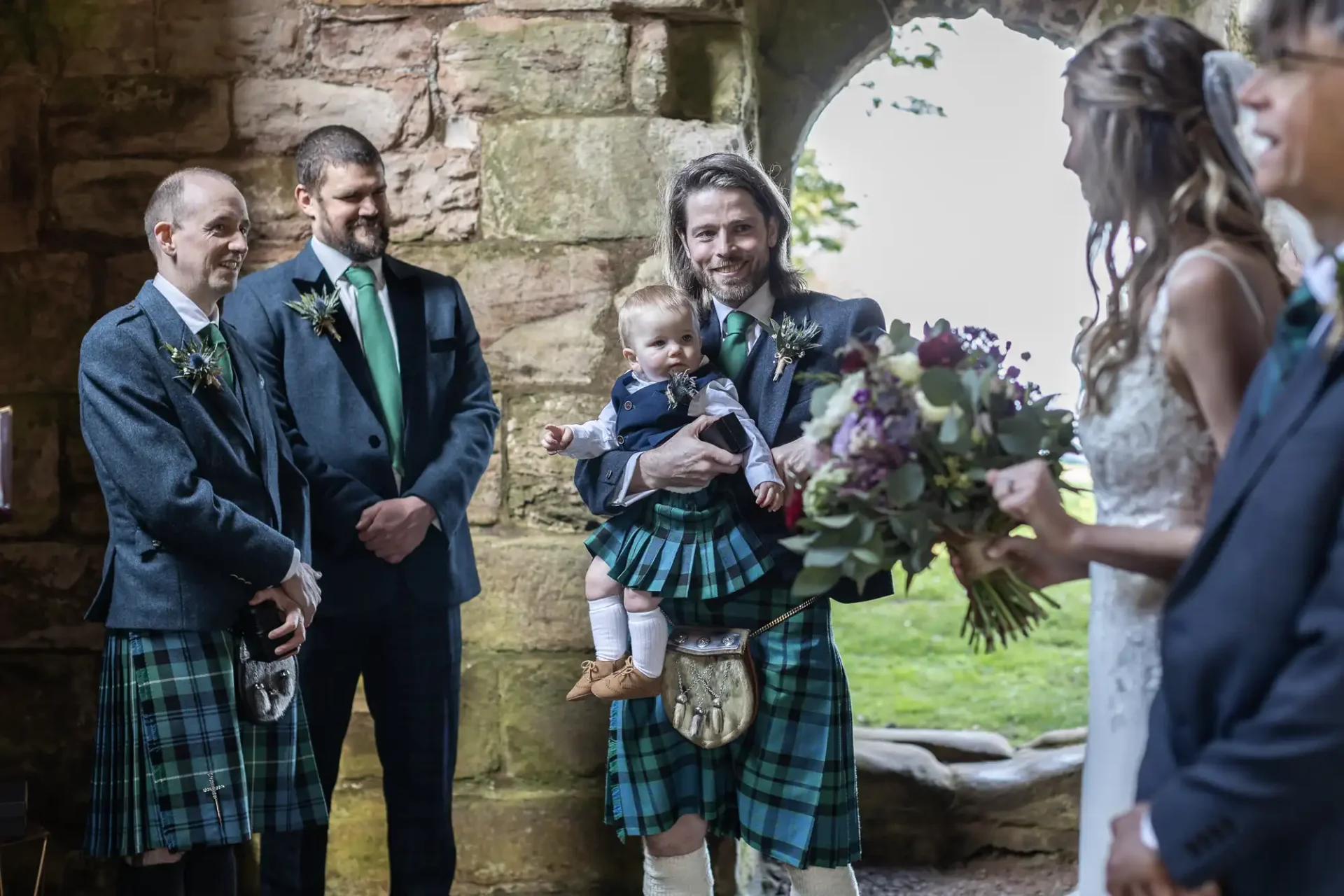 A wedding ceremony where three men in kilts, one holding a toddler, are standing beside a woman in a white dress holding a bouquet. They are inside a stone building with an archway.