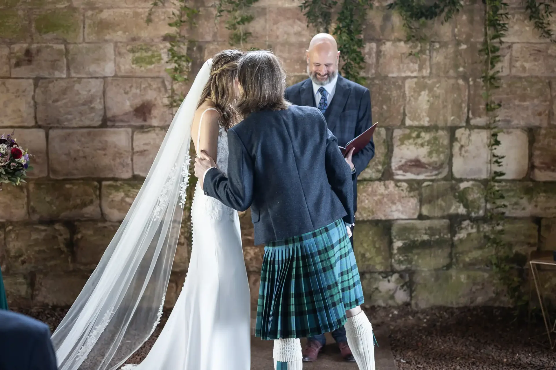 A bride in a white dress kisses a man in a kilt, while an officiant stands behind them, in front of a stone wall decorated with greenery.