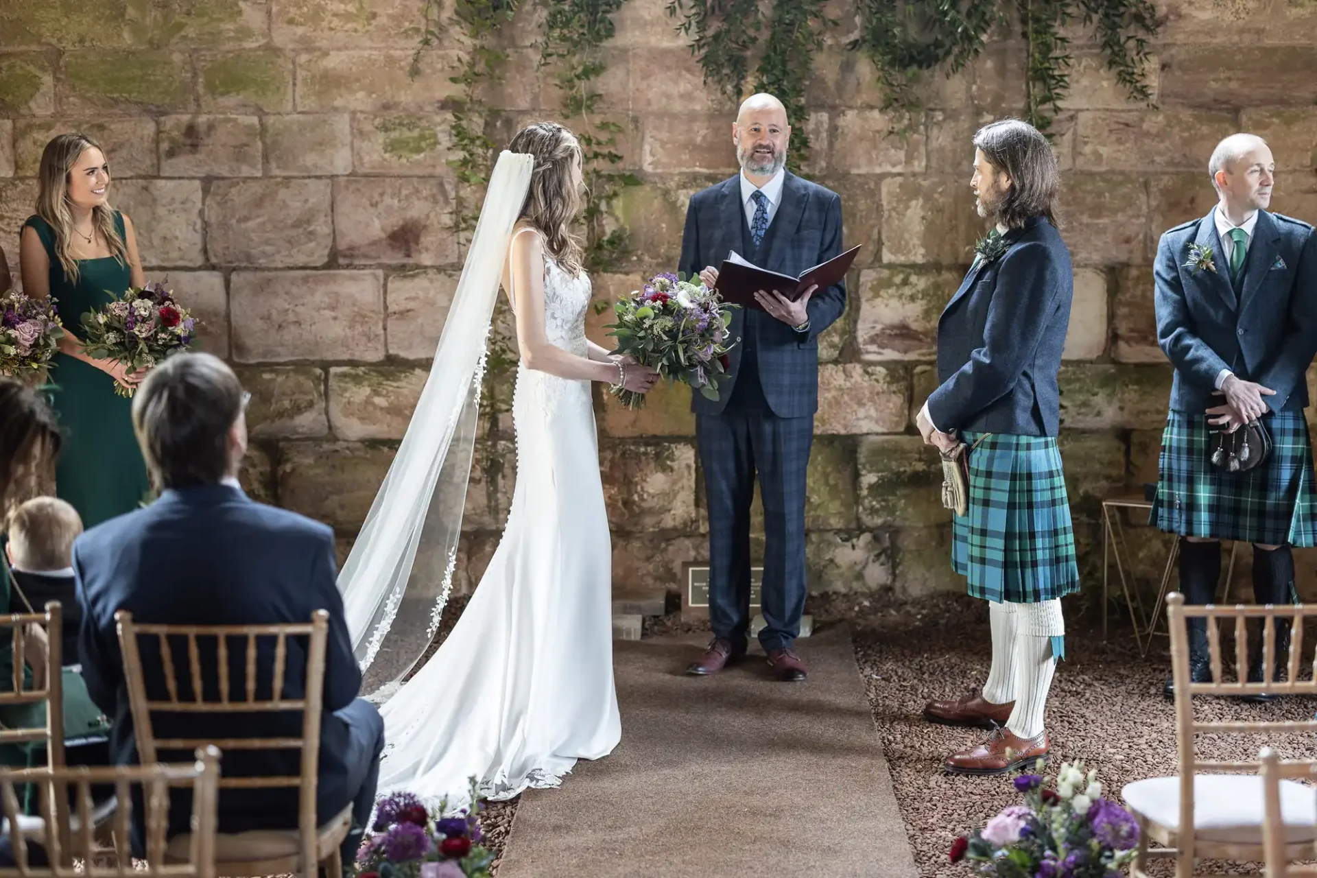 A wedding ceremony takes place with a bride in a white dress and veil and a groom in a kilt, standing before an officiant. Bridesmaids and guests are seated and observing the event in a rustic setting.